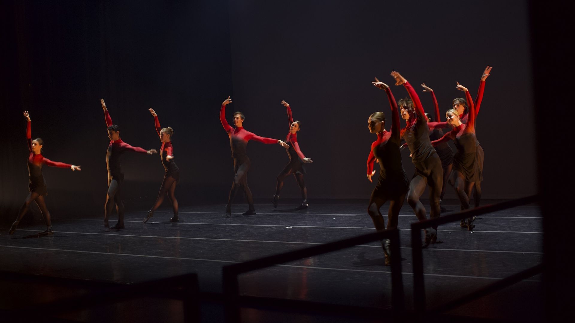 Group of dancers in red and black costumes perform on a dark stage, arms raised in synchronized, arcing motions.