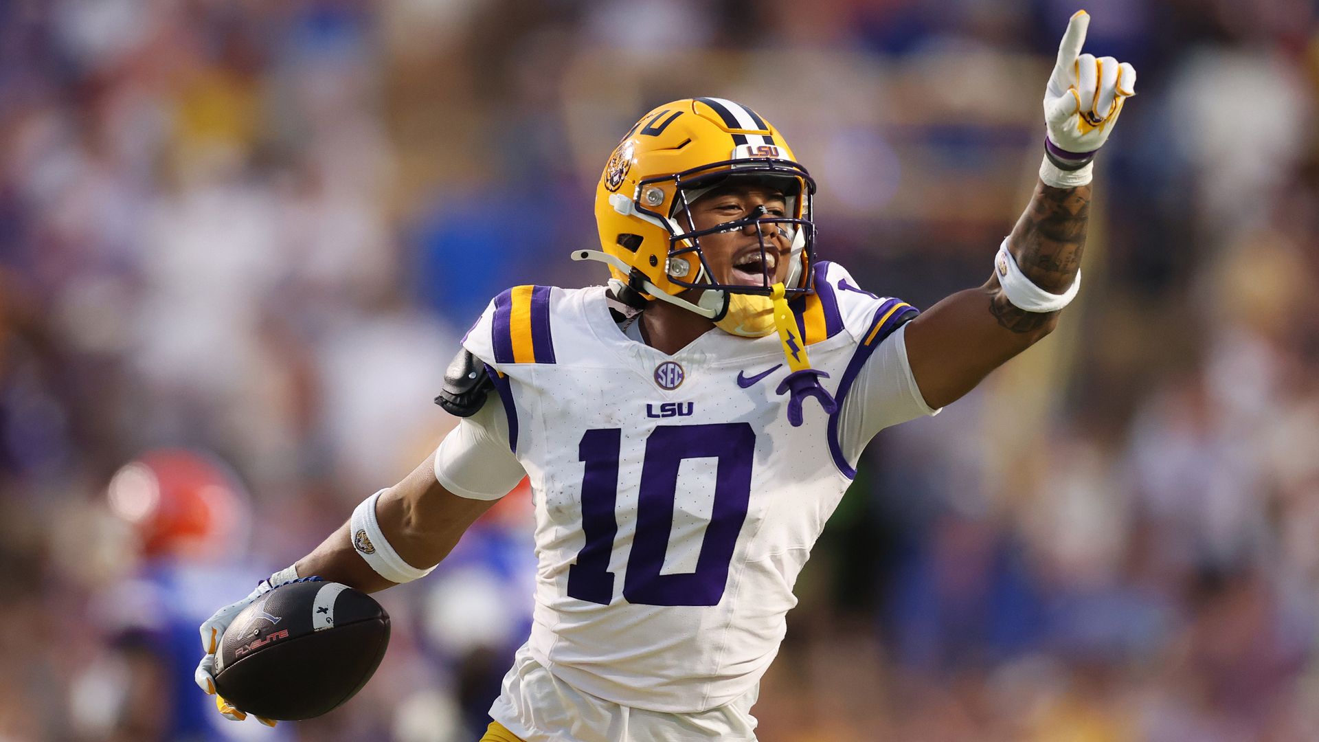 An LSU football player, wearing the No. 10 jersey, holds a finger into the air and the football with his other hand.