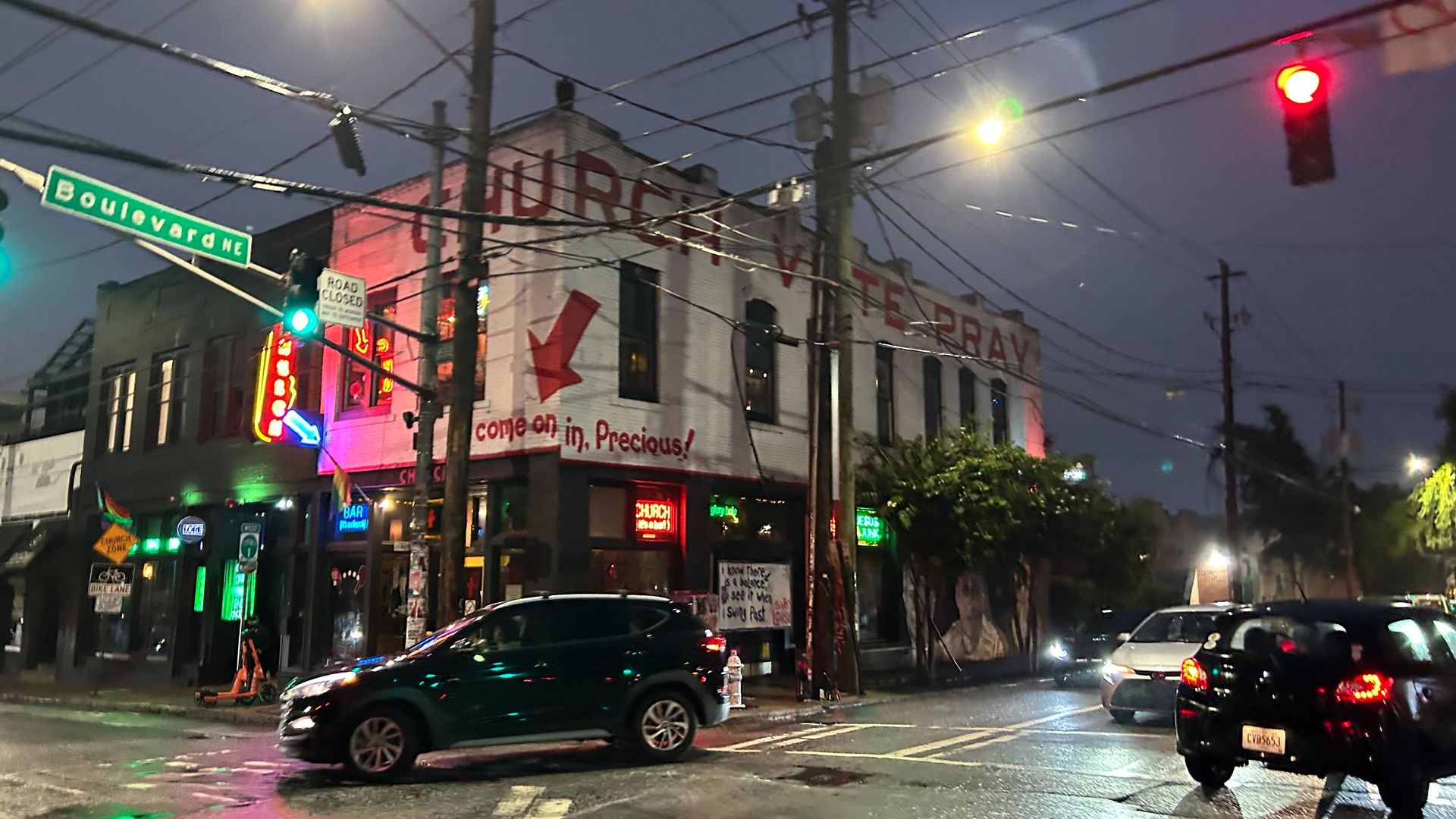 Night street scene at an intersection with traffic lights, cars, and a white two-story building with red lettering saying "CHURCH V TERRY, come on in, Precious!" and neon signs.