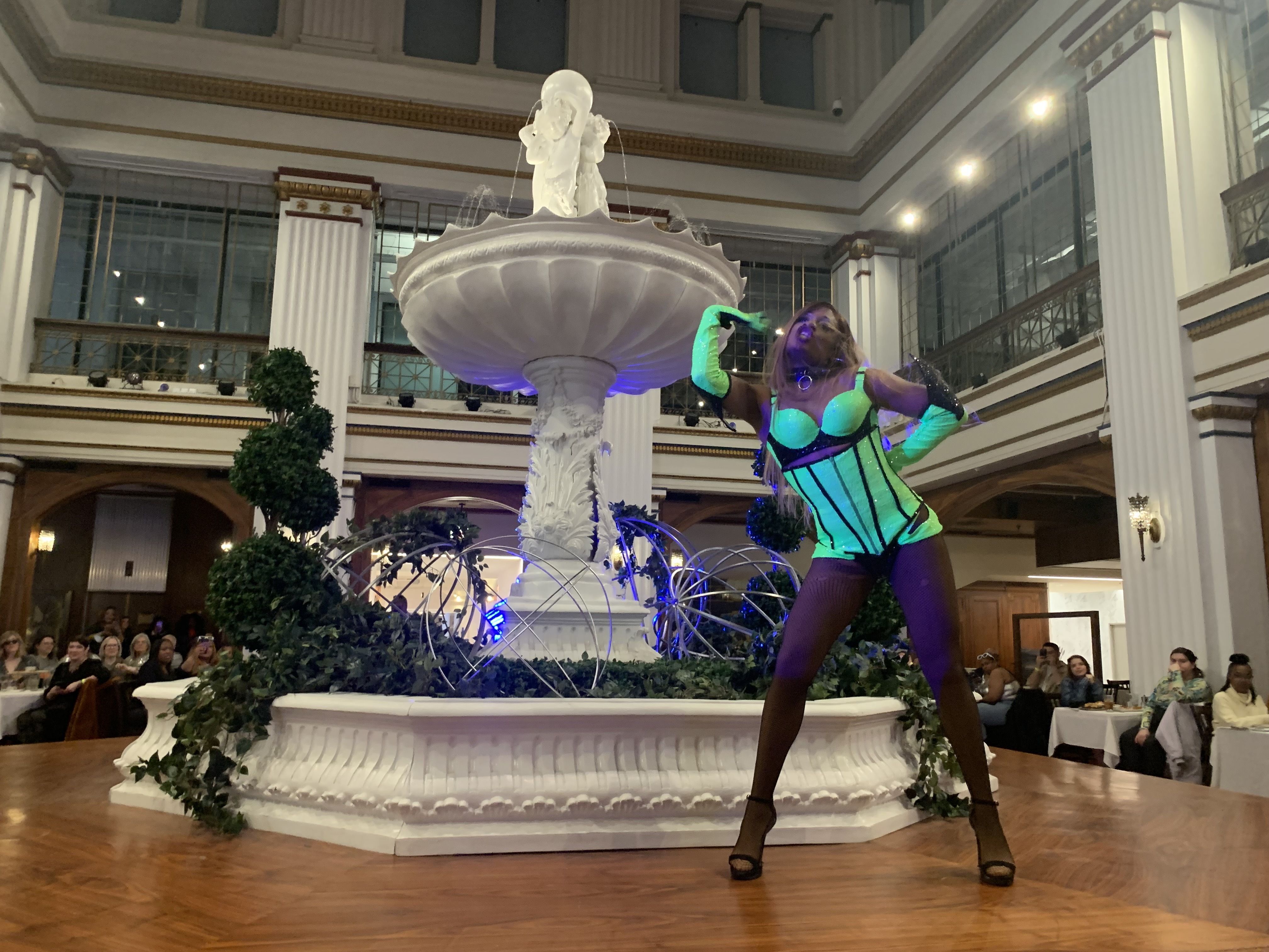 A drag performer in a bright green outfit dances in front of the large white fountain inside the Walnut Room.