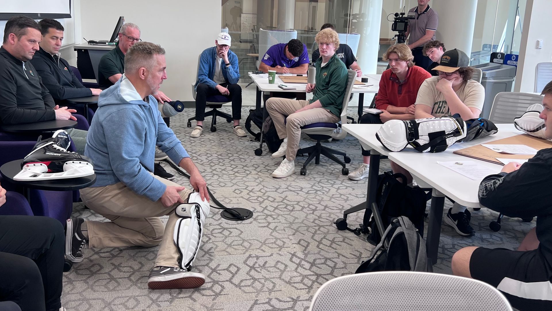 Group of people in a bright office conference room sit in a circle around a man in a blue hoodie kneeling with goalie pads; others watch as a cameraman films in the back.