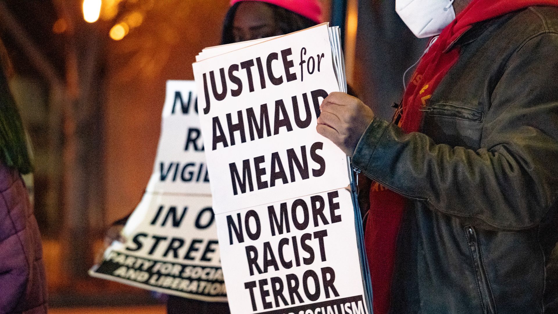 Photo of two people holding signs that say "Justice for Ahmaud Arbery means no more racist terror" at an outdoor vigil