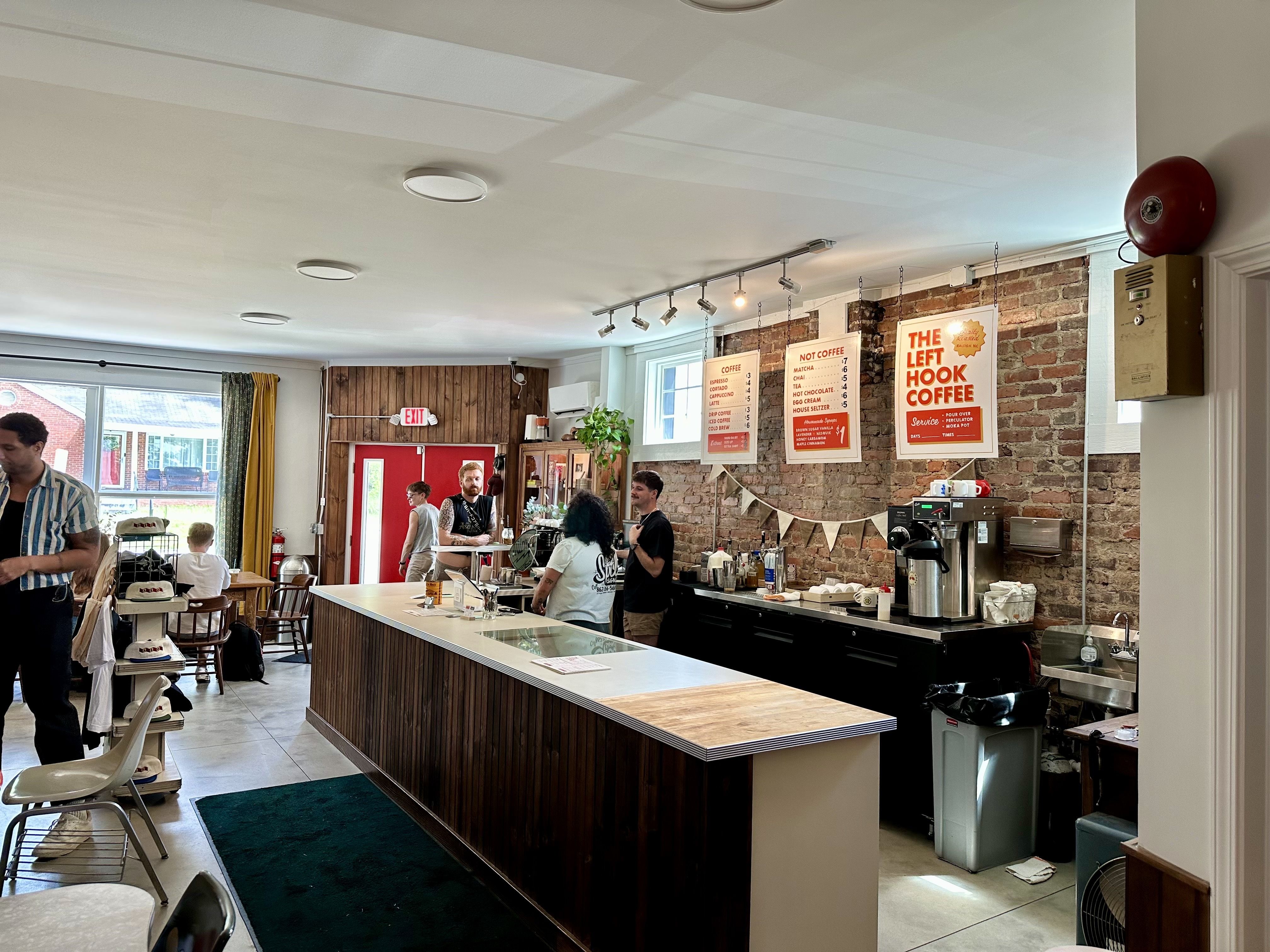 Cafe interior with a brick wall, a long wooden counter, and track lighting. Several customers stand at the bar under posters, including "The Left Hook Coffee". Large windows have yellow and green curtains.