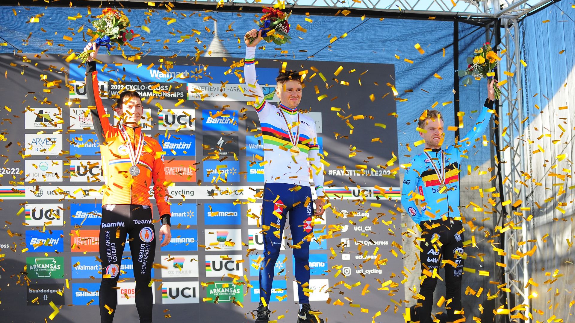 Three cyclists stand at the winner's podium holding flowers.