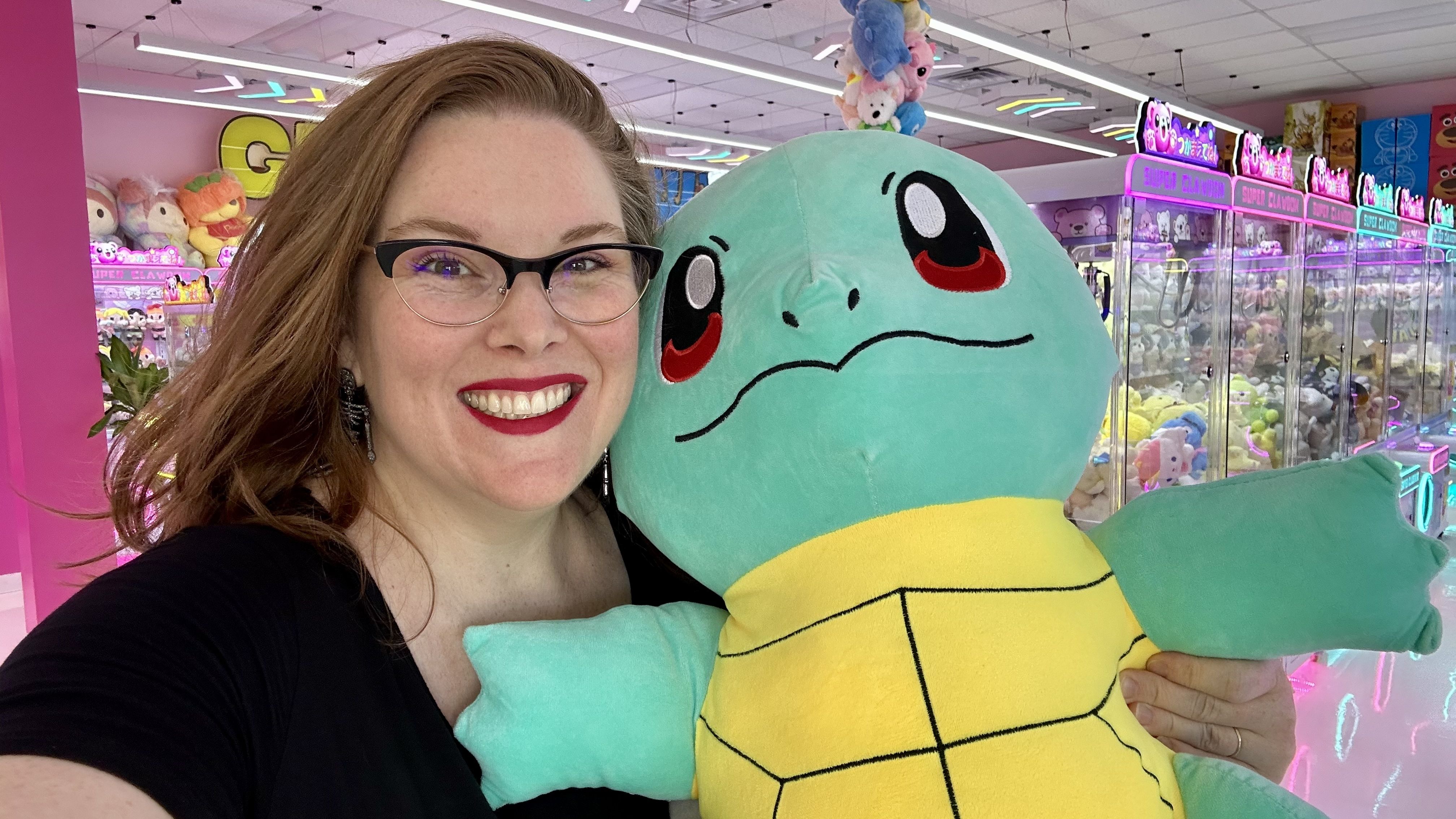 Smiling woman with glasses and red lipstick holding a large blue and yellow Squirtle plush in a brightly lit arcade filled with claw machines and colorful stuffed toys.