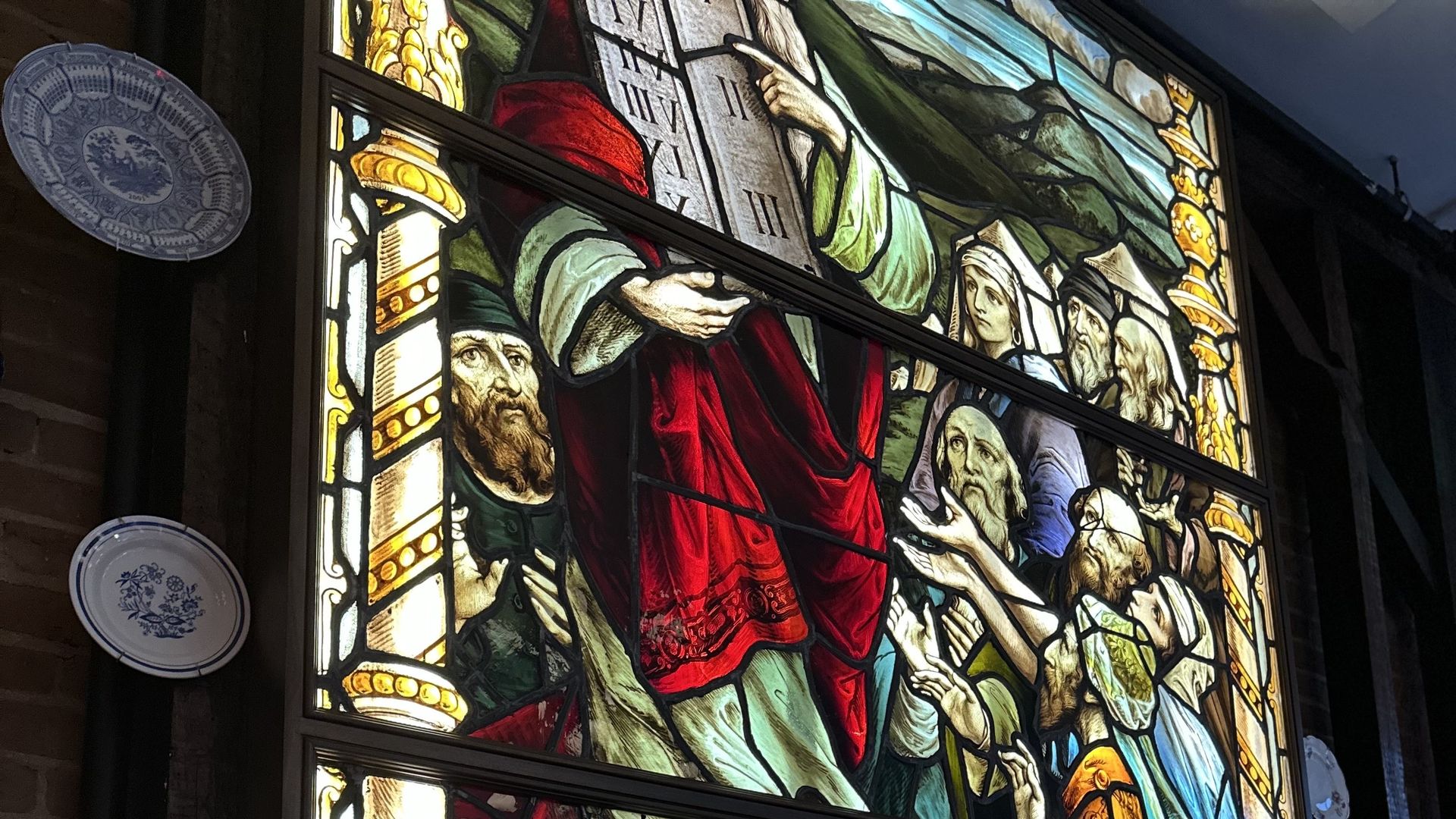 Colorful stained-glass window depicting a biblical scene with a central red-robed figure surrounded by people; ornate gold trim, stone wall exterior, two decorative plates on left, red chairs and a table in front.