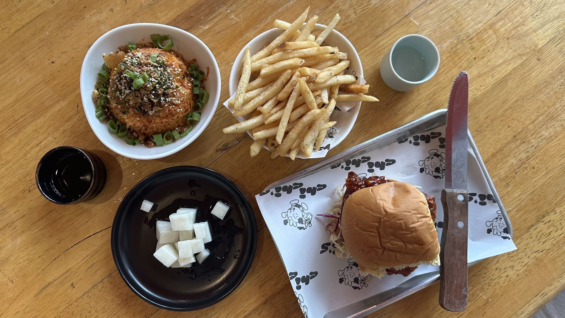 A tabletop is viewed from above with a chicken sandwich, fries, and a bowl of kimchi fried rice.