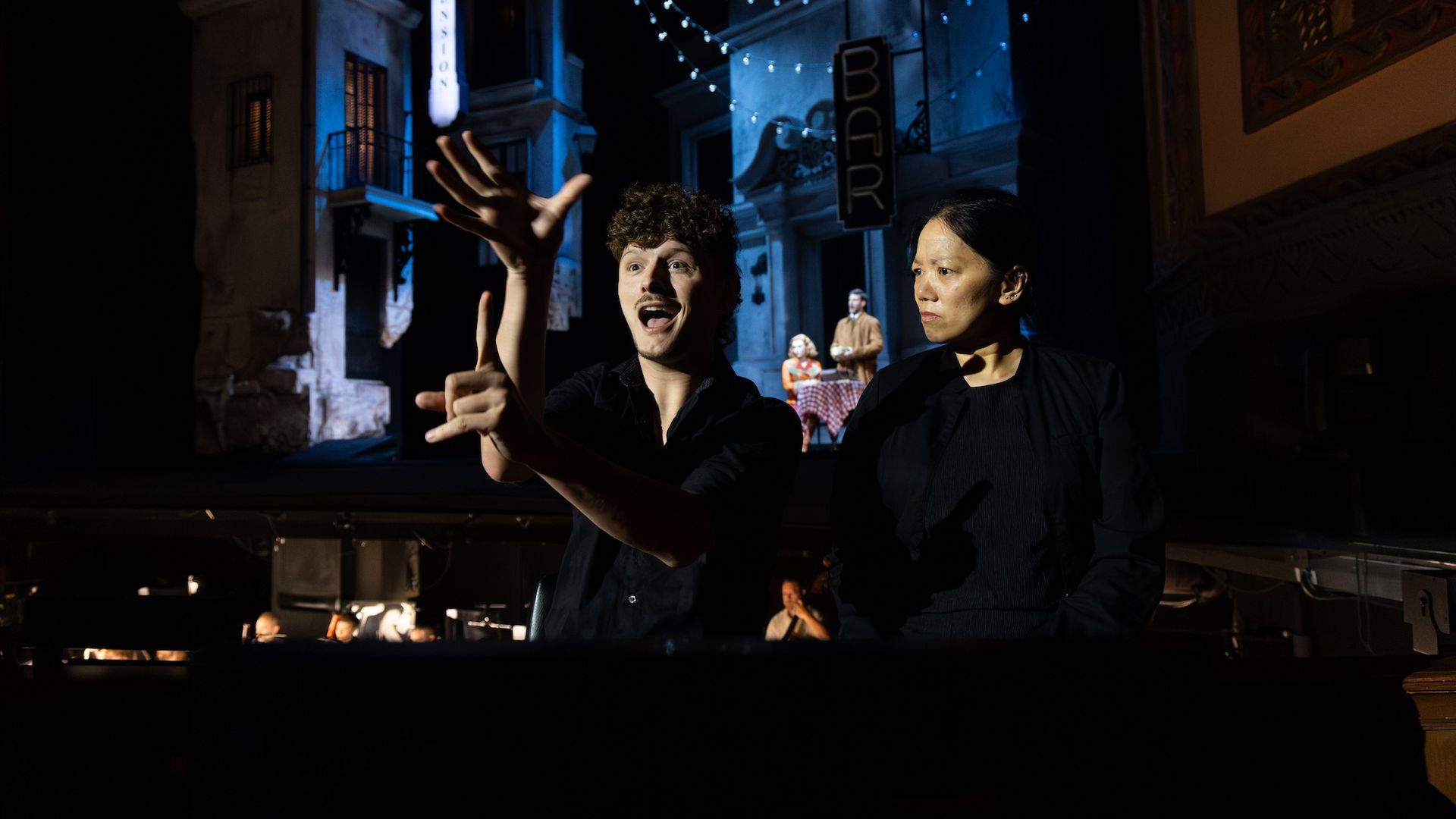 Two actors in the foreground on a dark stage; a curly-haired man gestures excitedly to the audience while a serious woman stands beside him. Blue-lit street backdrop with string lights behind them.