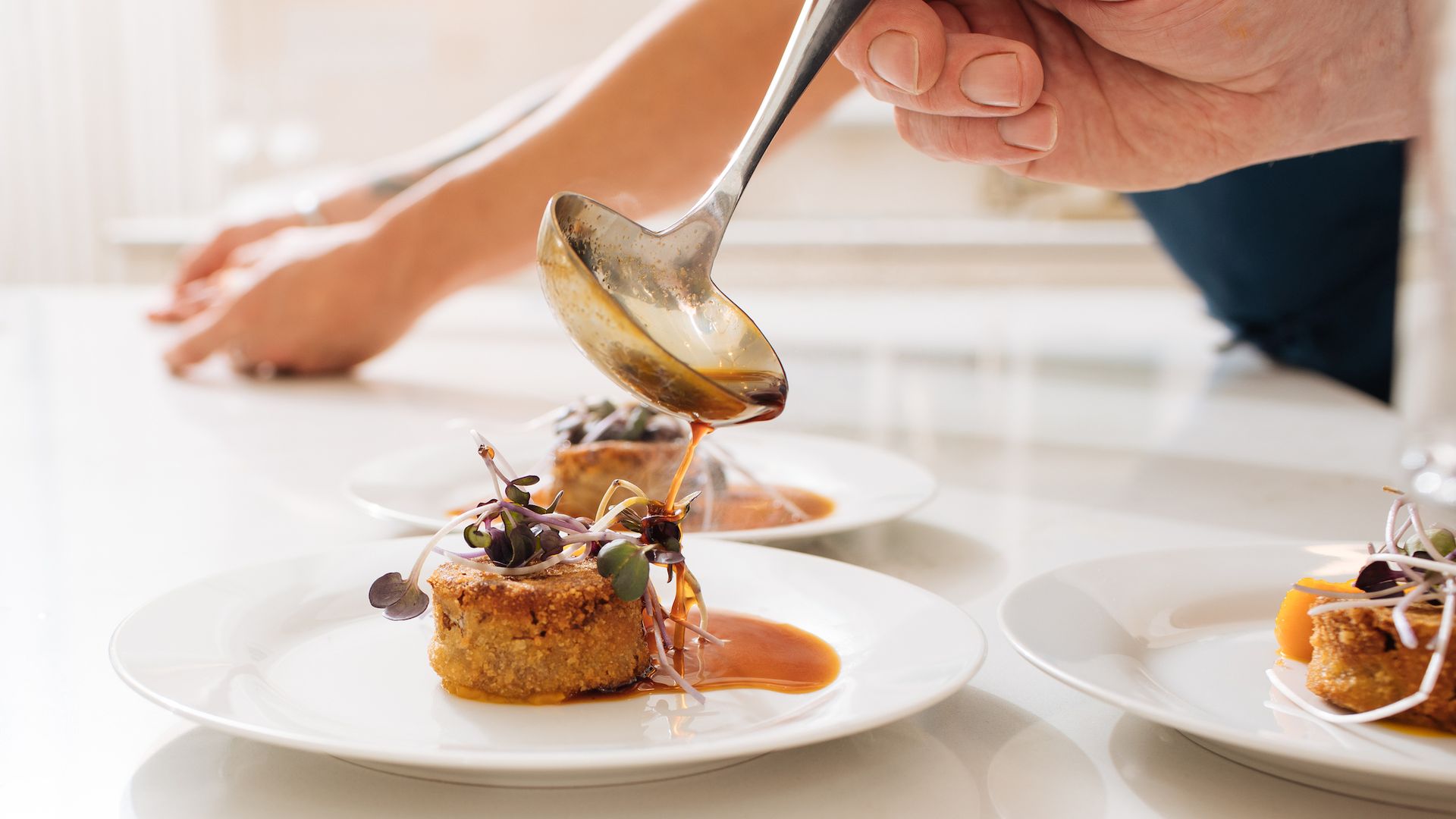 A hand uses a ladle to pour brown sauce over a plated gourmet dish with microgreens on a white plate, set on a white countertop with two similar dishes in the background.