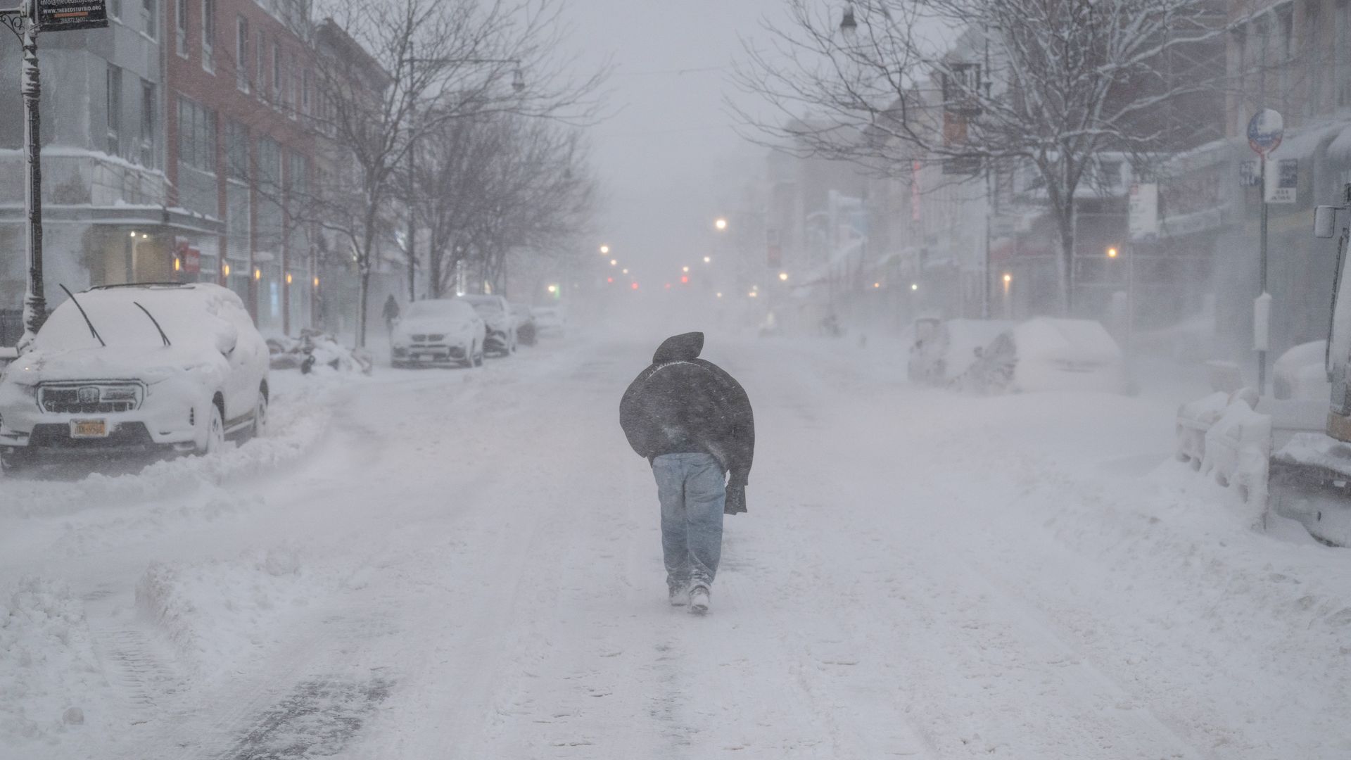 A person wearing blue jeans and a jacket with the hood up walks down a snow-covered street lined with cars blanketed with snow.