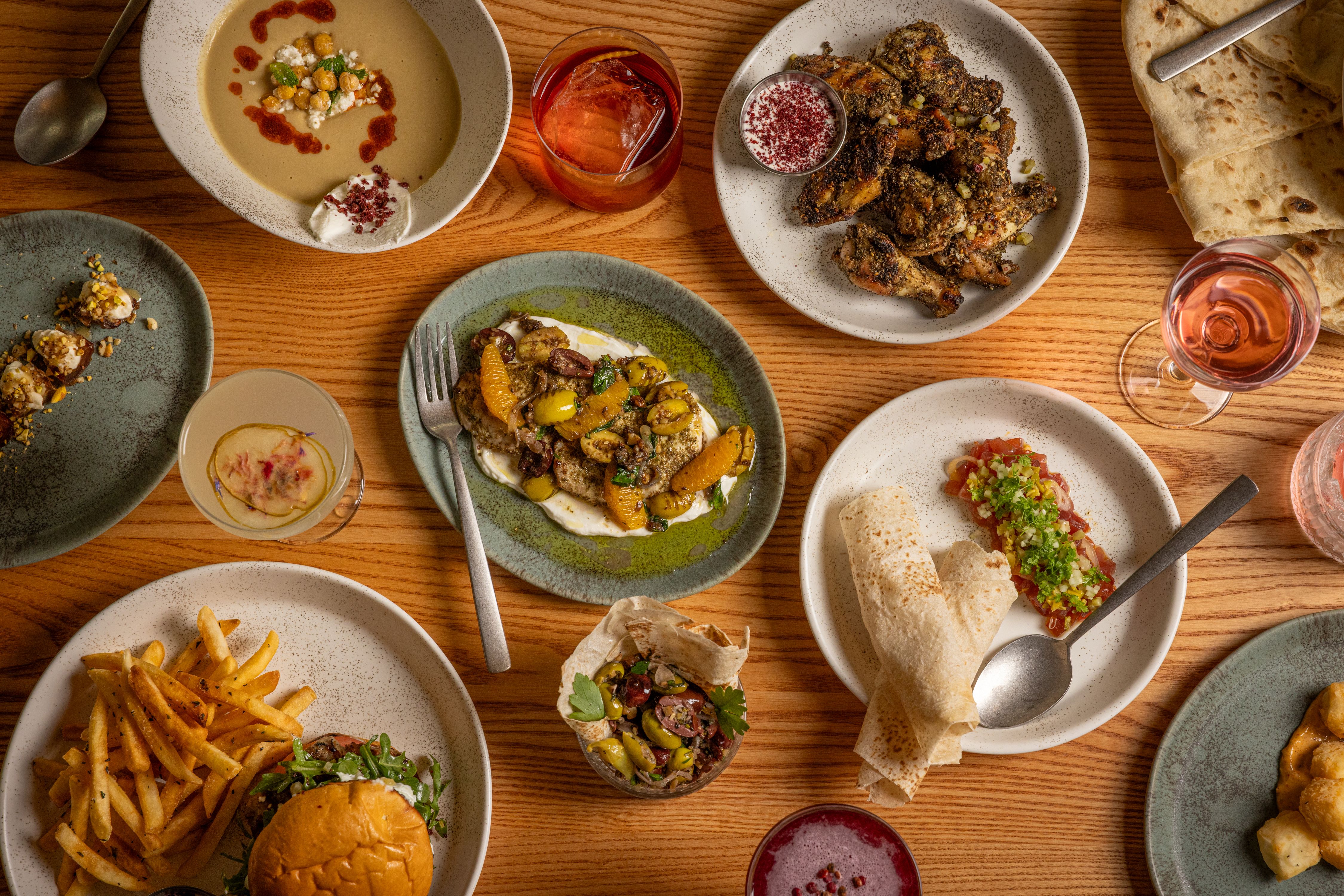 Wooden table with plates of fried chicken wings, soup with chickpeas, a burger with fries, citrus and herb dish, olives in tortilla, flatbread with tomato salad, and drinks including a pink wine.