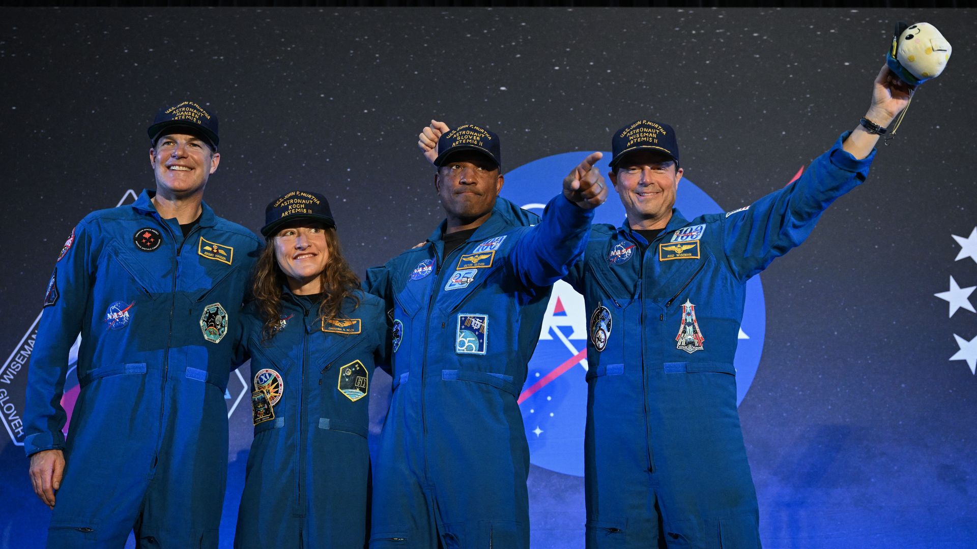 Four astronauts in blue jumpsuits stand on a stage with a starry backdrop, smiling for a group photo. Patches on their suits; one raises a small yellow-and-white plush toy.