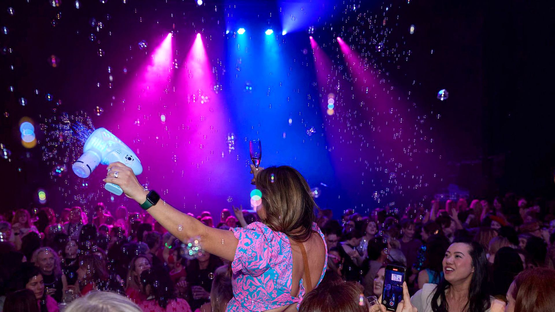 Crowd at a party; a woman in a pink patterned dress faces away, raises a blue bubble machine and a champagne flute as pink and blue stage lights shine and bubbles fill the air.