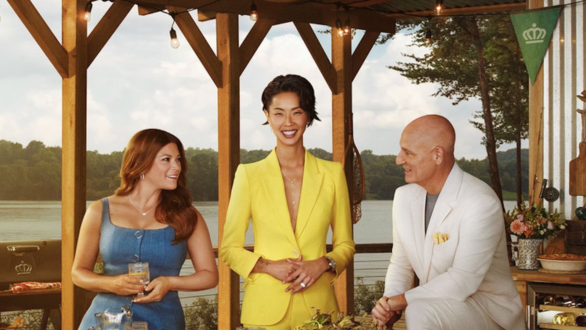 Three people smiling and standing around a table filled with various grilled and fresh foods on a wooden deck overlooking a lake, with a wooden ceiling and string lights above.