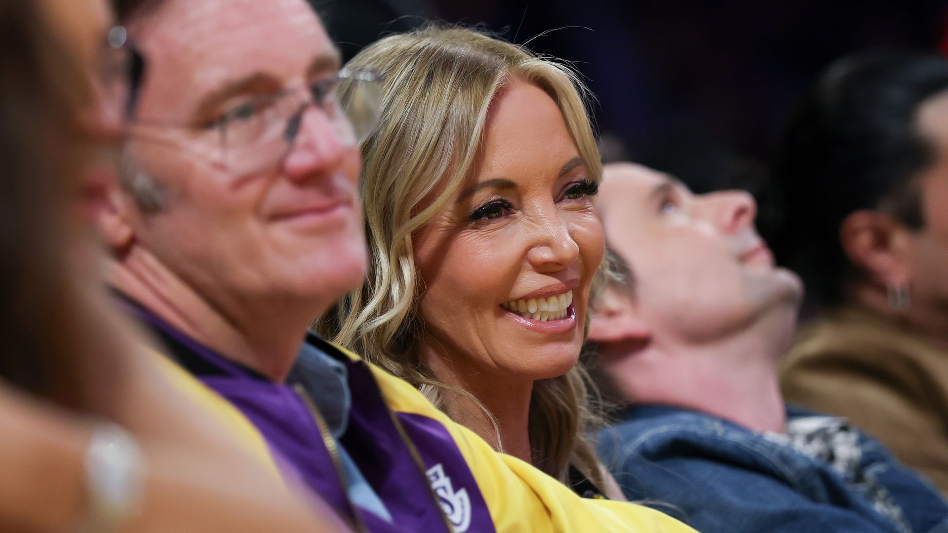 Close-up of a smiling blonde person in a yellow shirt and purple lanyard seated in an audience, with other attendees blurred in the background.