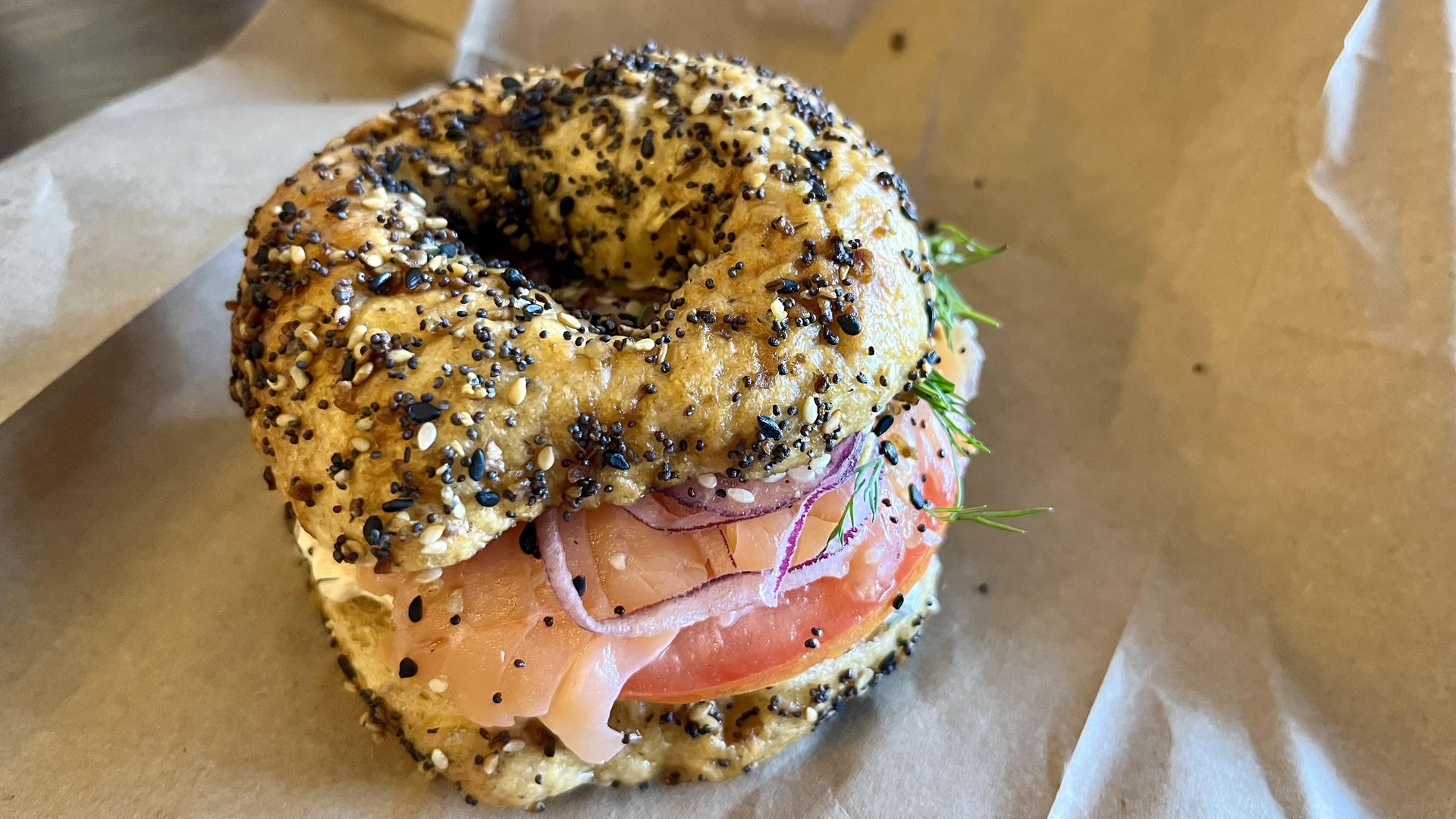 Everything bagel sandwich with smoked salmon, red onion slices, tomato, and dill on brown paper.