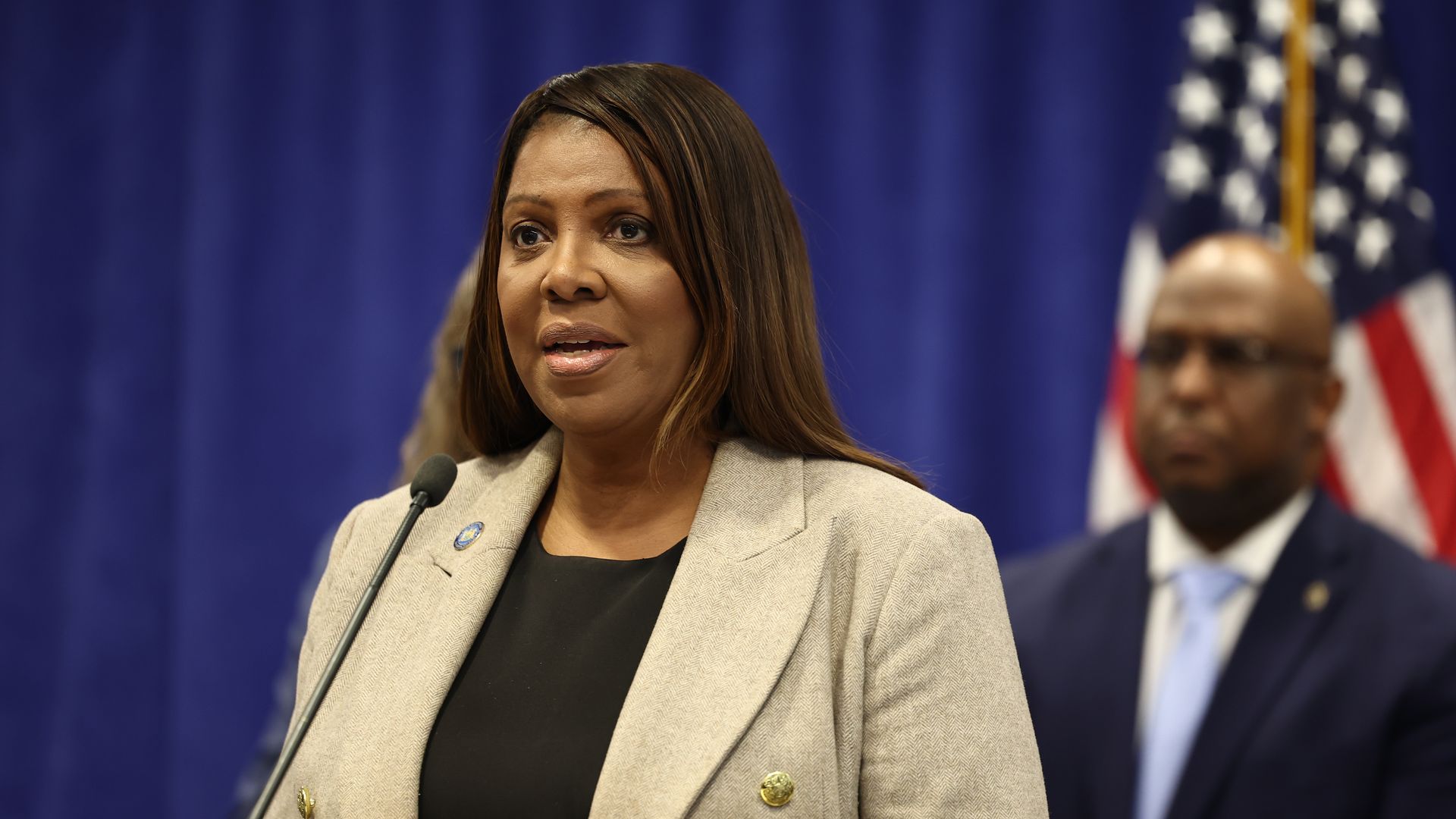 NY Attorney General Letitia James, wearing a beige coat and black top, stares ahead while speaking into a black mic. A blue curtain and blurred man can be seen in the background.