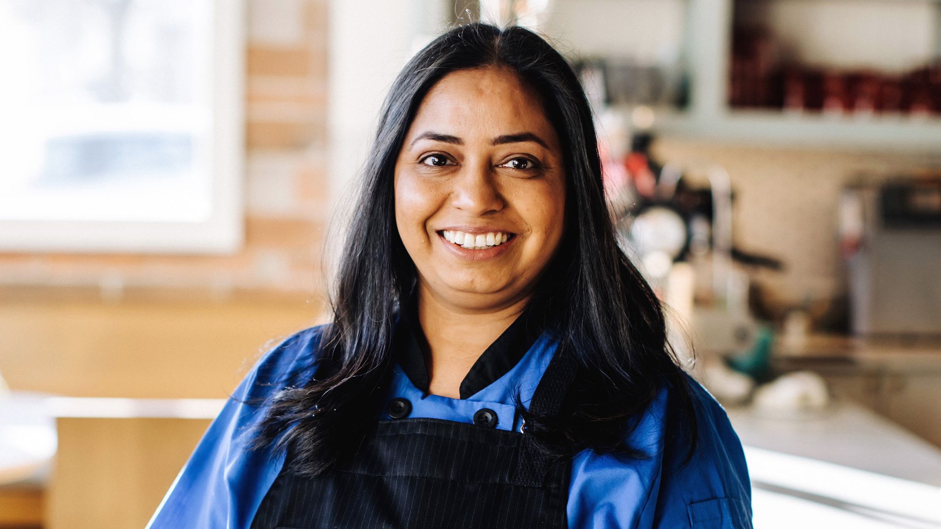 Jyotiee Kistner wears a blue shirt and black apron and stands in a kitchen
