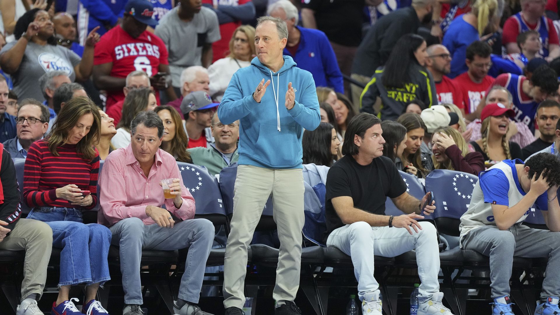 Josh Harris is standing and clapping courtside on the sidelines in between seated fans at a packed 76ers game.