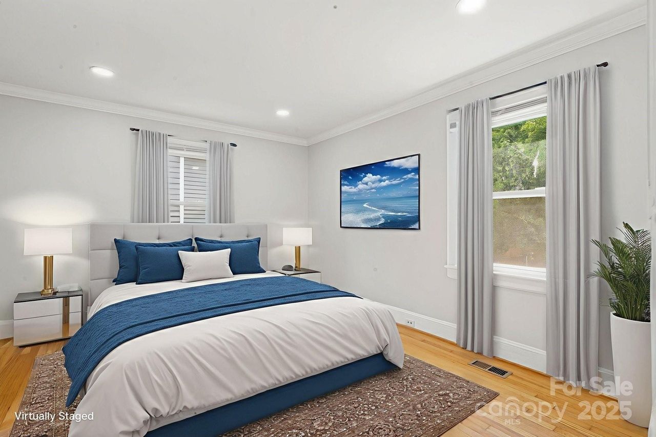 Bright bedroom with white walls, gray curtains, king bed with blue and white bedding, two gold and white lamps on mirrored nightstands, wall art of ocean waves, wooden floor, and indoor plant.