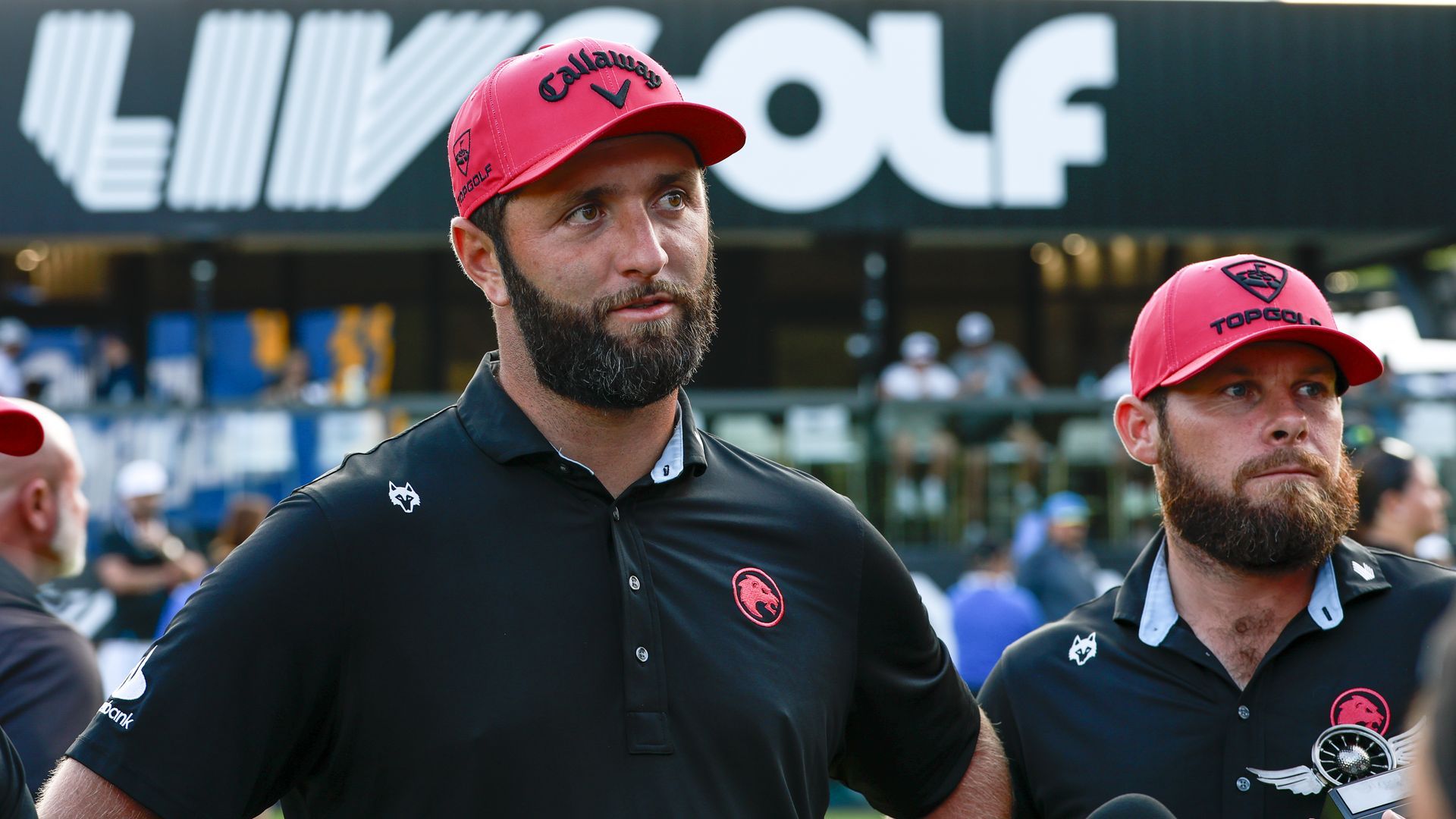 Two bearded men in black polo shirts and bright red caps stand outdoors at a LIV Golf event, with a large sign behind them and a blurred crowd in the background.
