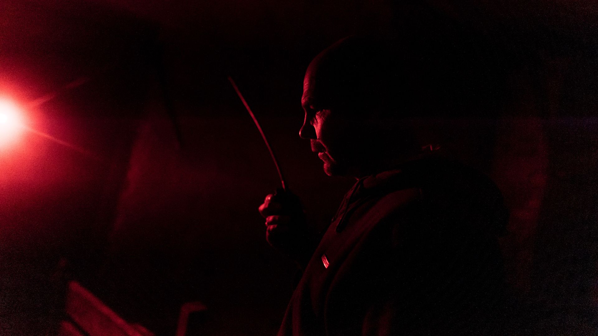 A Ukrainian soldier stands in a dark room, gripping a radio. The only source of light is to the left of the frame; it is glowing red.