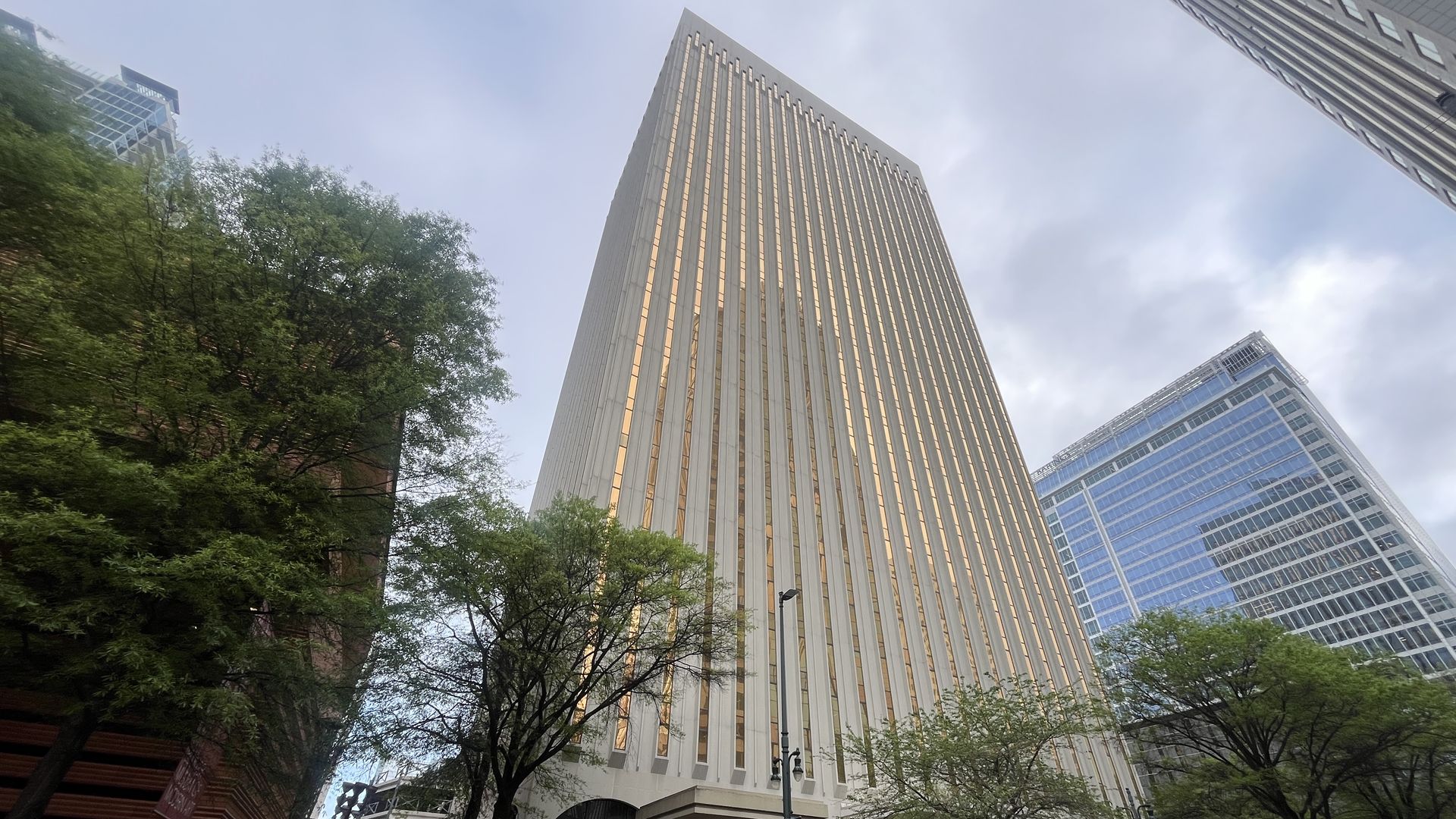Tall city street featuring a cream-colored skyscraper with vertical windows, framed by green trees. A blue glass tower stands to the right, with a U-Haul truck in the foreground and a cloudy sky above.