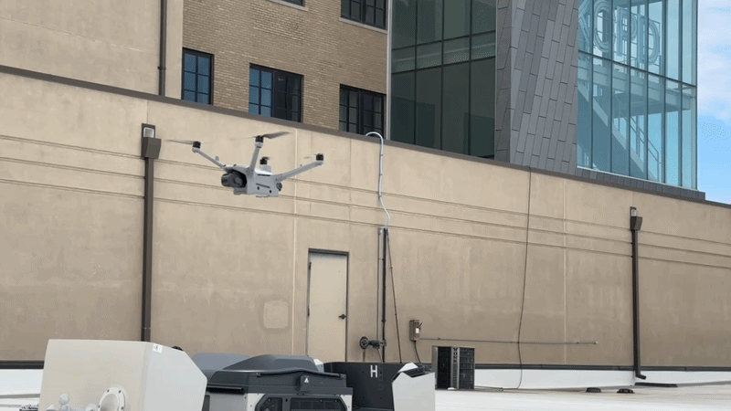A gray drone with four arms and a camera flies near a beige building wall and a pane of glass windows in the back displaying the letters "KCPD."