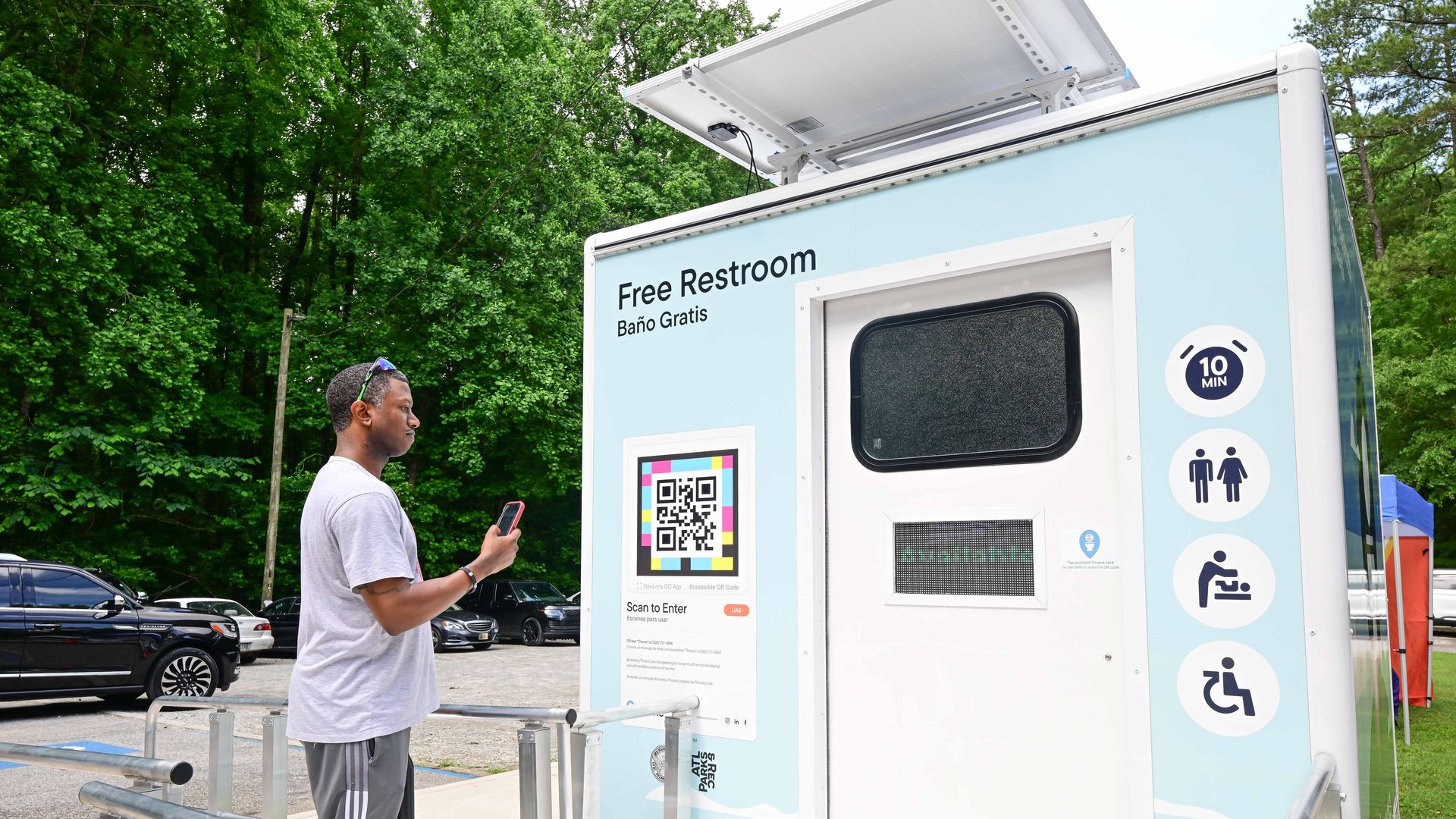 An outdoor view of a light-blue restroom trailer with a large 'Free Restroom' sign and 'Baño Gratis'. A man in a gray shirt stands holding a phone near the trailer railing, with a QR code and accessibility icons visible.