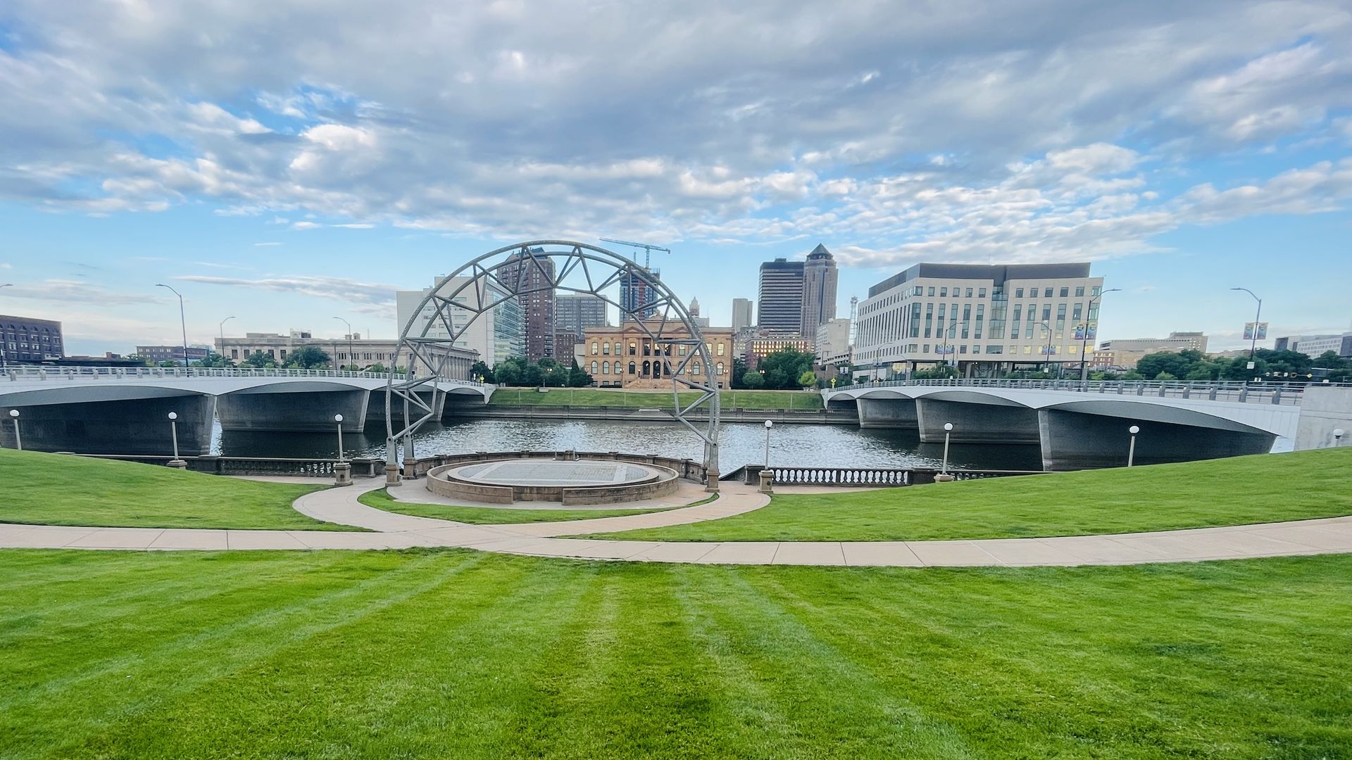 City skyline with modern buildings across a river; a large metal arch sculpture stands in the water between two bridges, in a green park with a circular plaza in the foreground, under a cloudy blue sky.