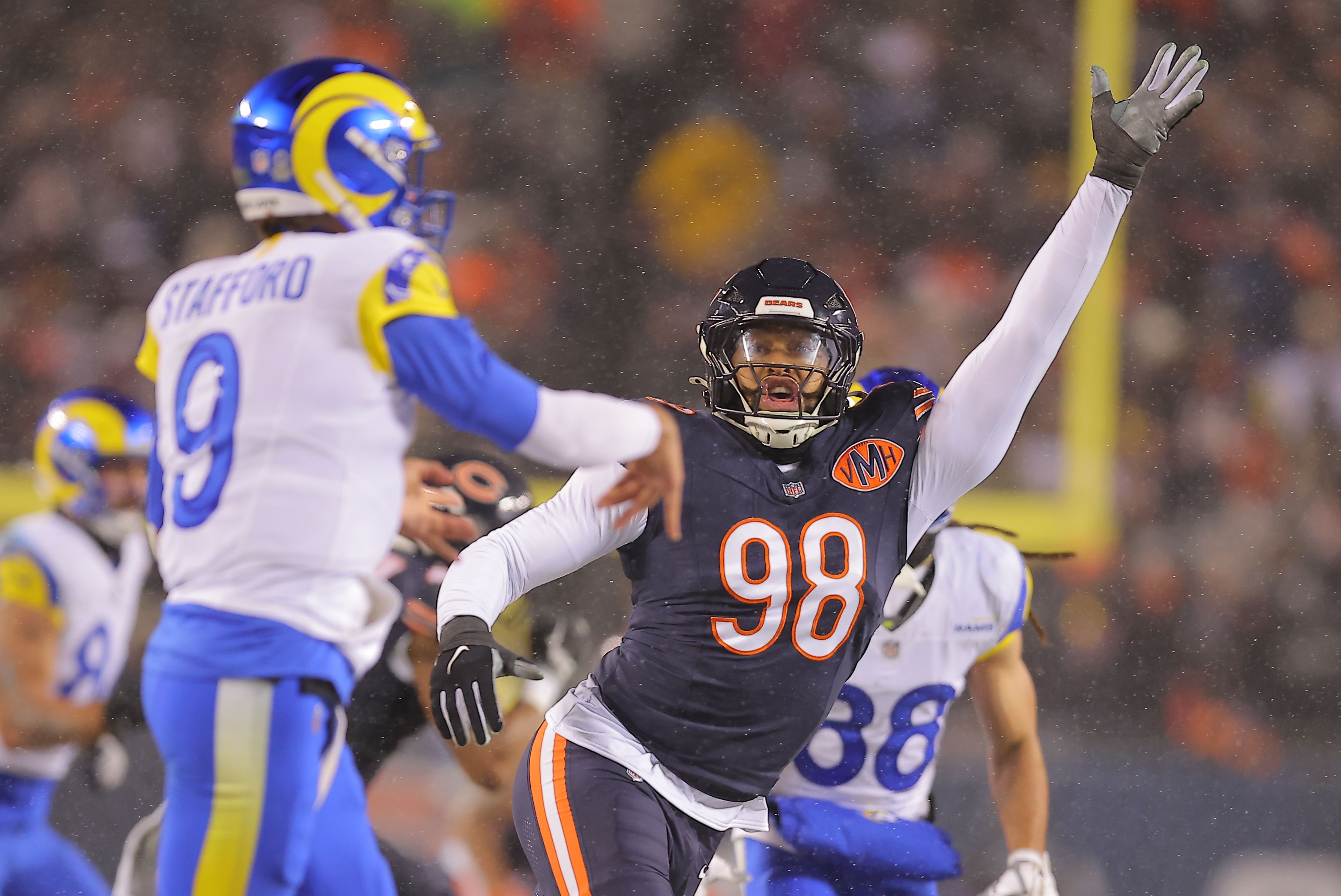 Rainy football scene: a Chicago Bears player in navy jersey with orange 98 raises his gloved right hand as a blue-and-yellow opponent approaches, with a blurred crowd in the background.