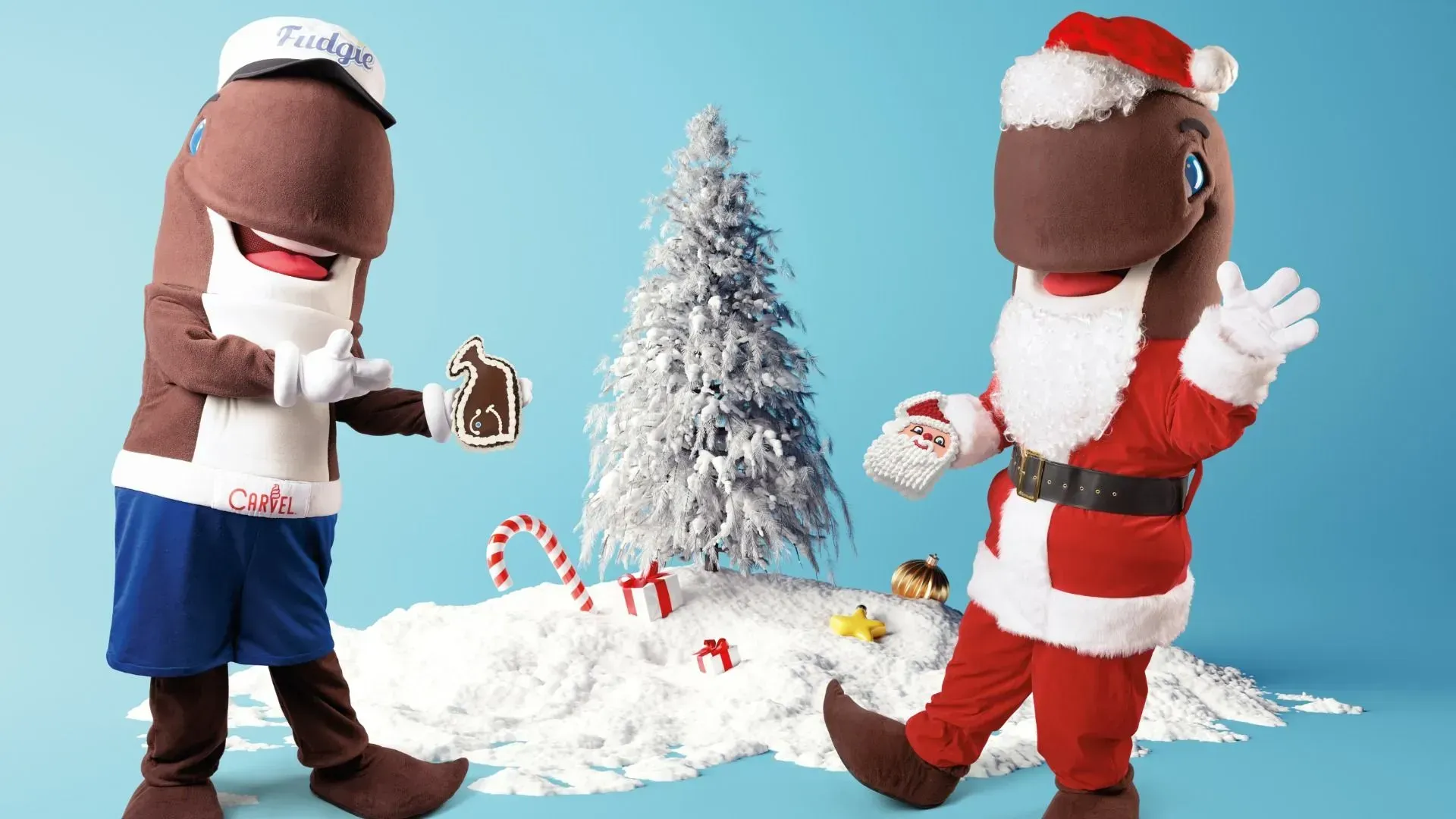 Two large plush mascots in a snowy blue scene: left wears a white cap labeled Fudgie and apron, holding a cookie; right is in a Santa suit with white beard, waving. Snowy ground, frosted tree, candy canes, gifts.