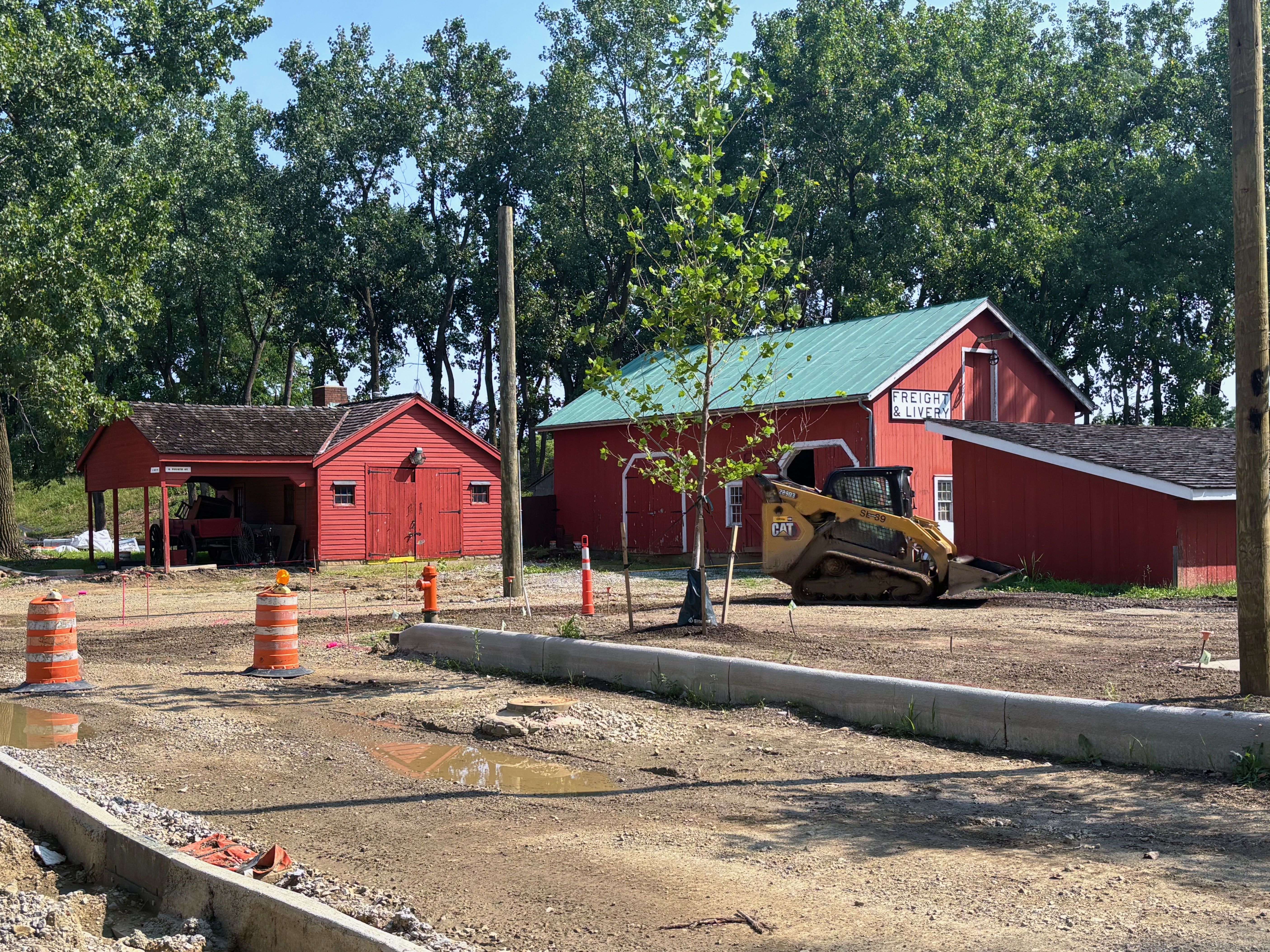 Red buildings with a green roof sit among tall green trees; a young sapling grows in front, while orange traffic cones and a CAT excavator mark a muddy construction site.