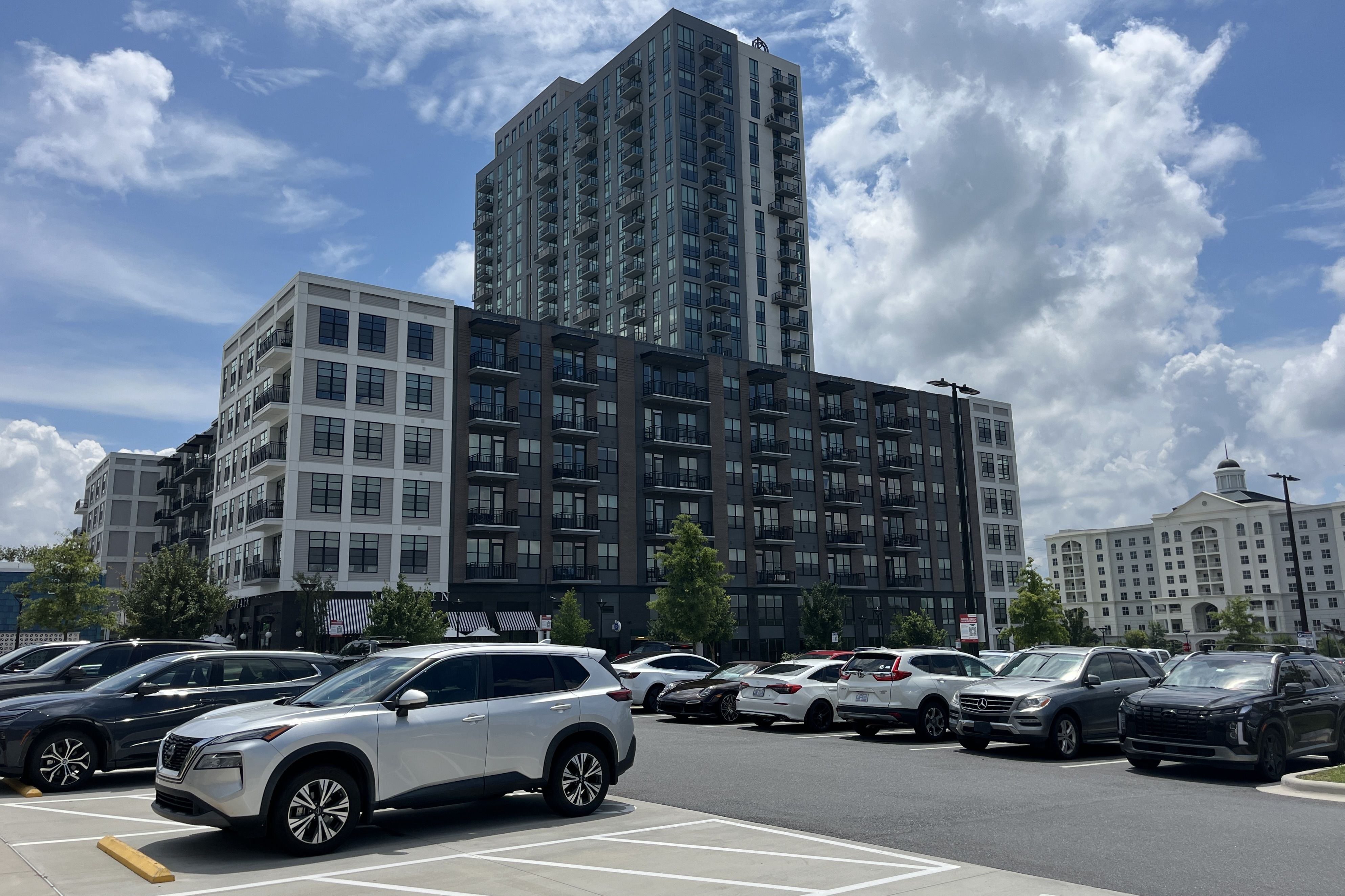 Modern apartment complex with a tall central tower and balconies, white and gray facade, under a blue sky with clouds; a busy parking lot filled with cars in the foreground.