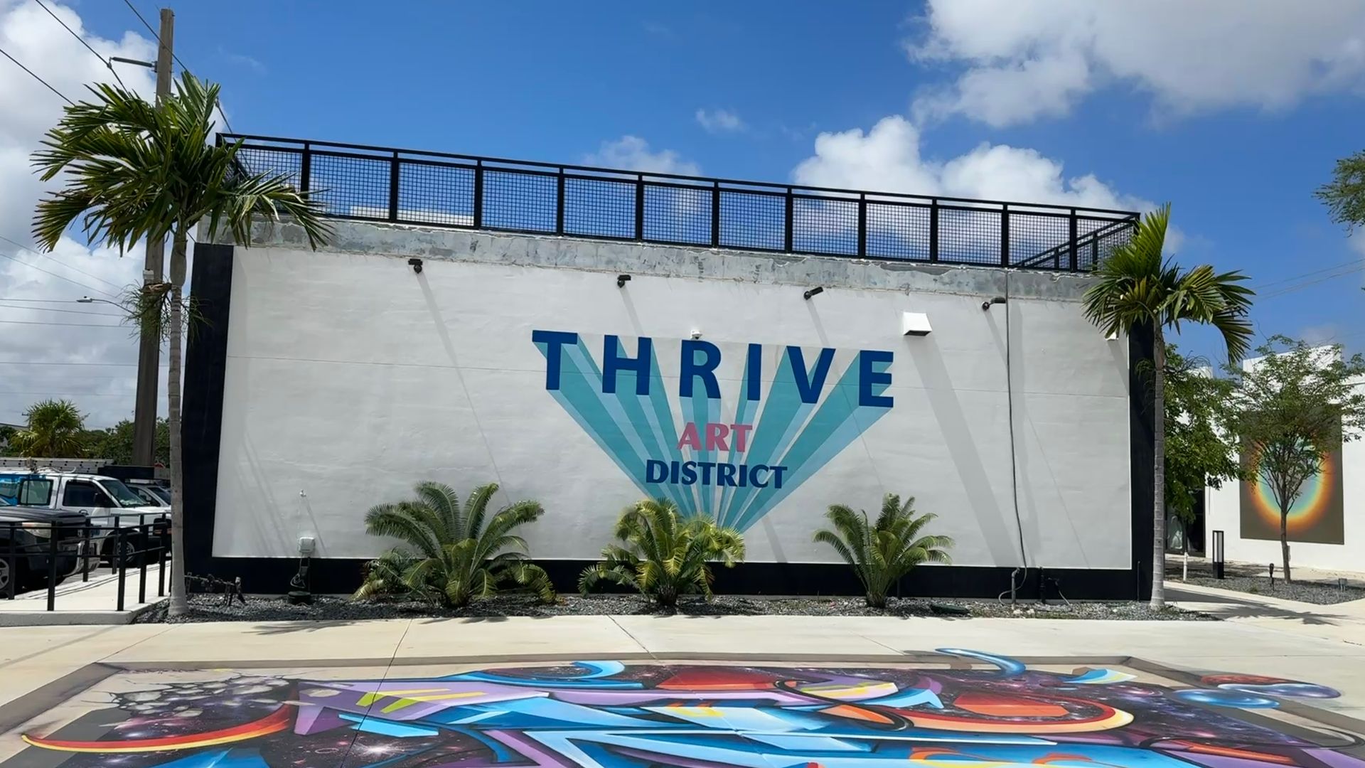 Bright mural on a white wall reading "THRIVE ART DISTRICT" with teal sunburst rays; colorful abstract art on the pavement in the foreground. Palm trees, blue sky with clouds, and a black railing along the roof.