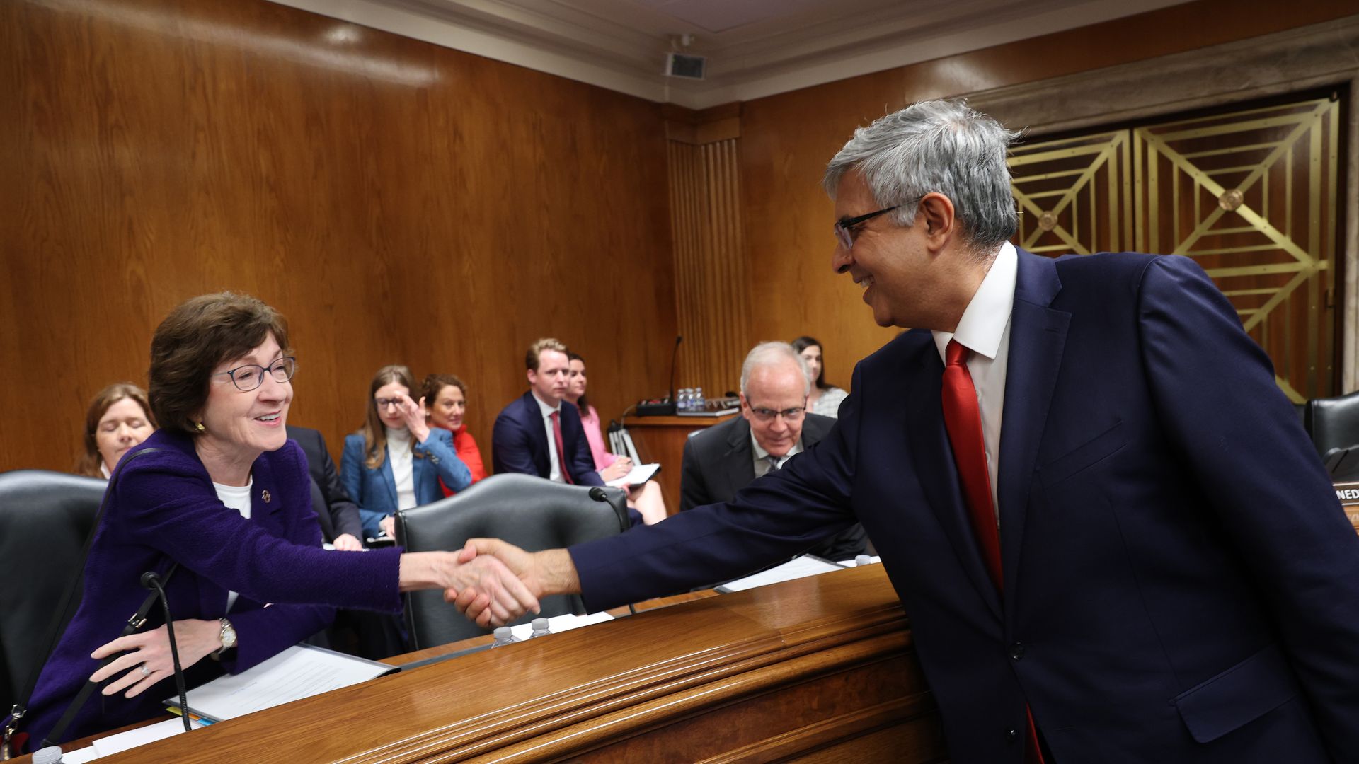 Jay Bhattacharya shakes Susan Collins' hand over a wooden desk in a Senate hearing room.
