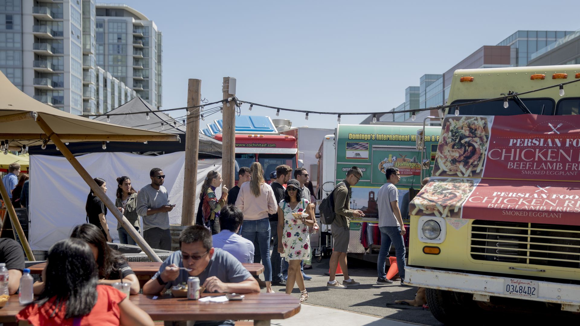 Outdoor food market with string lights, tents, and colorful food trucks. People walk and sit at wooden tables as modern apartment buildings rise in the background.