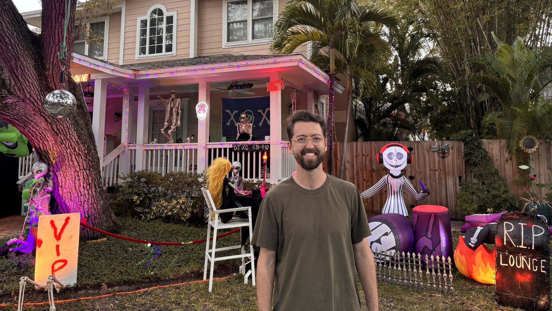 Pink, two-story house decorated for Halloween with purple lights and skeleton figures in the yard; a smiling man in a green shirt stands in the foreground, with a large tree trunk on the left.