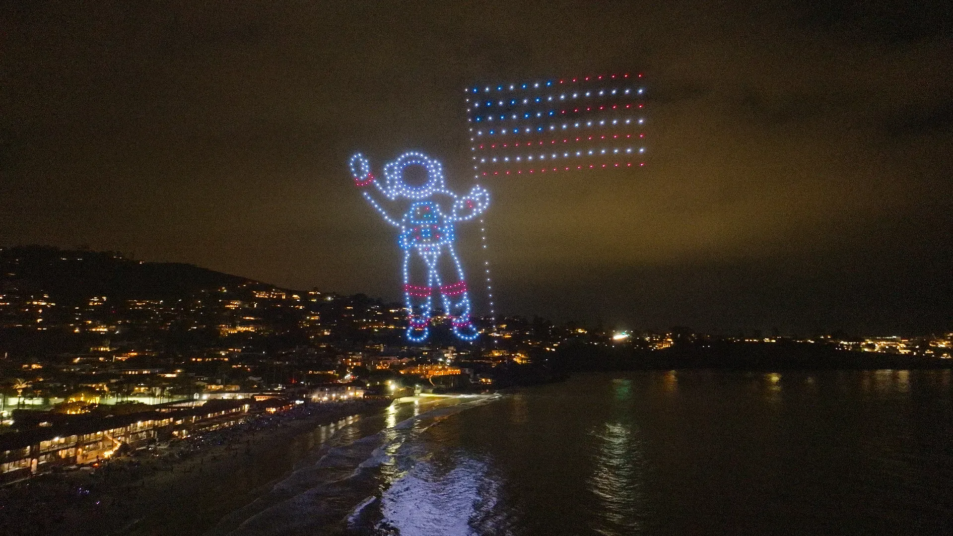 Night view of a giant LED-lit astronaut figure hovering over a coastal town. Blue-white outline with red accents, arm raised; a dotted US flag extends to the right, city lights and water below.