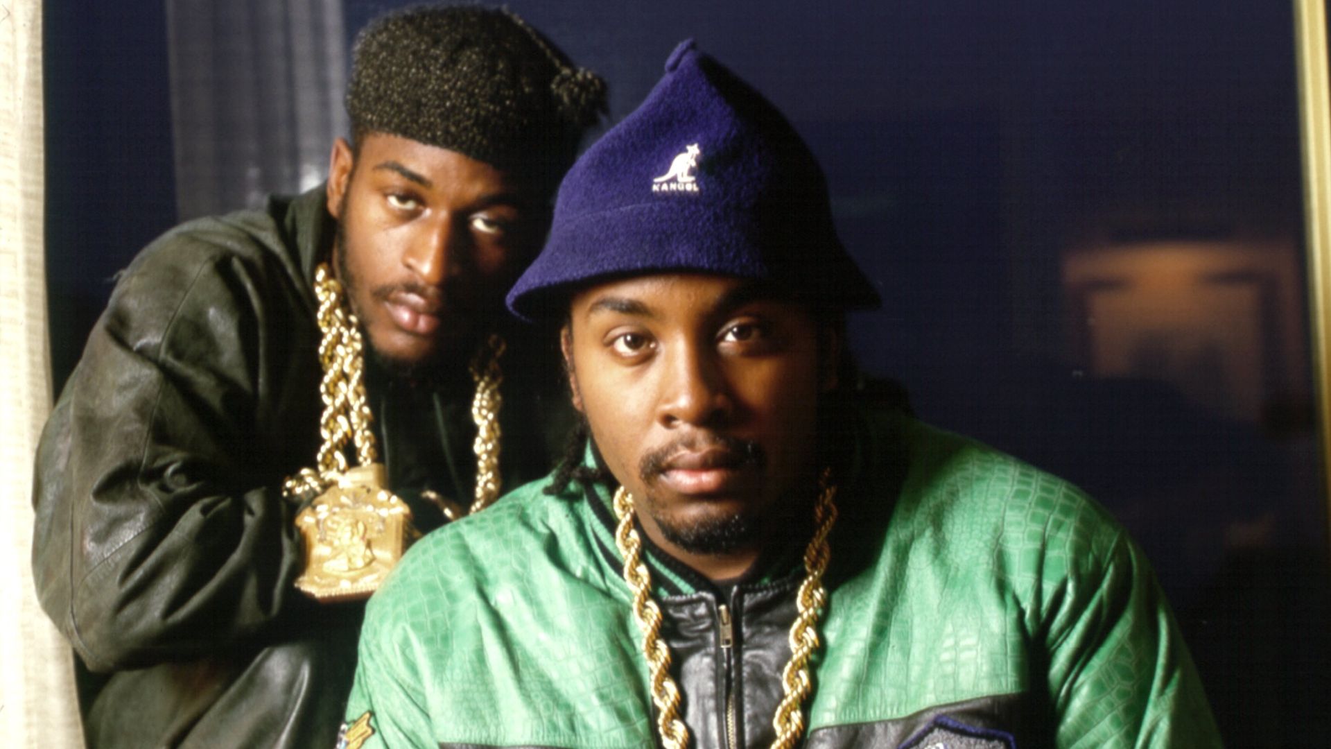 Two men wearing gold chains and rings, one in a green and black jacket with a blue Kangol hat, the other in a dark jacket and black beanie, posing indoors with serious expressions.