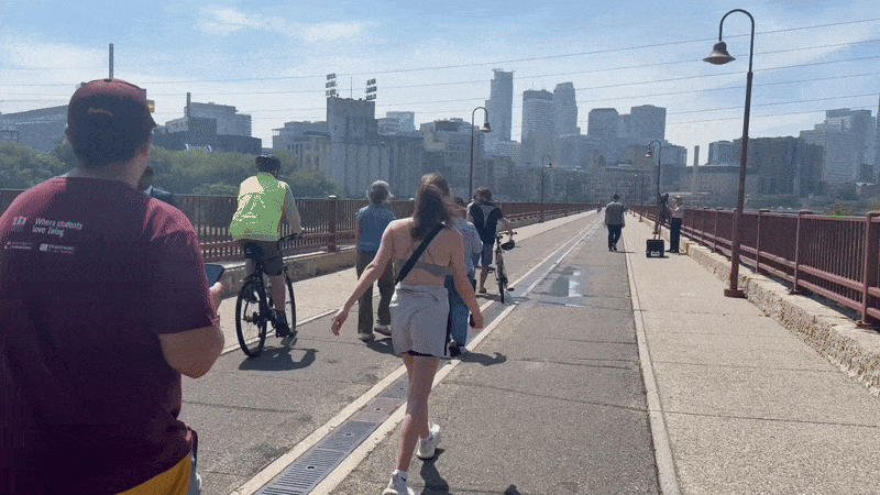 People walking and cycling on a sunny urban bridge with red railings and street lamps, city skyline visible in the background under a blue sky.