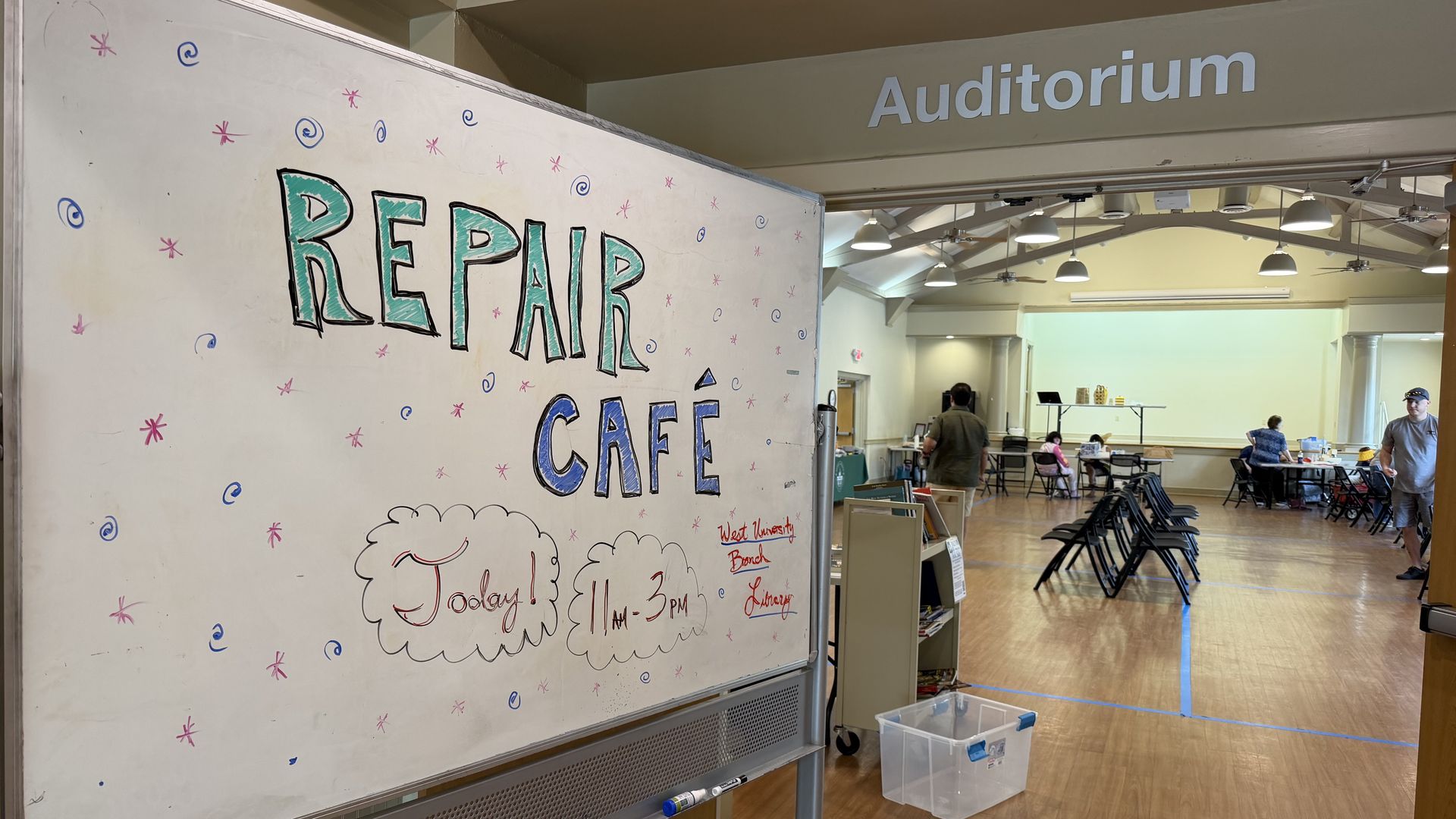 Whiteboard with colorful doodles reading REPAIR CAFE in teal letters; in the background an auditorium sign, ceiling lights, and people at tables in a bright community space.