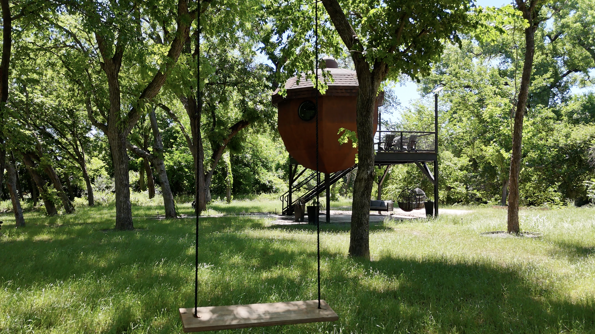 A photo of an acorn-shaped tree house on a platform in a wooded area. A swing hangs in front of it on a tree. 