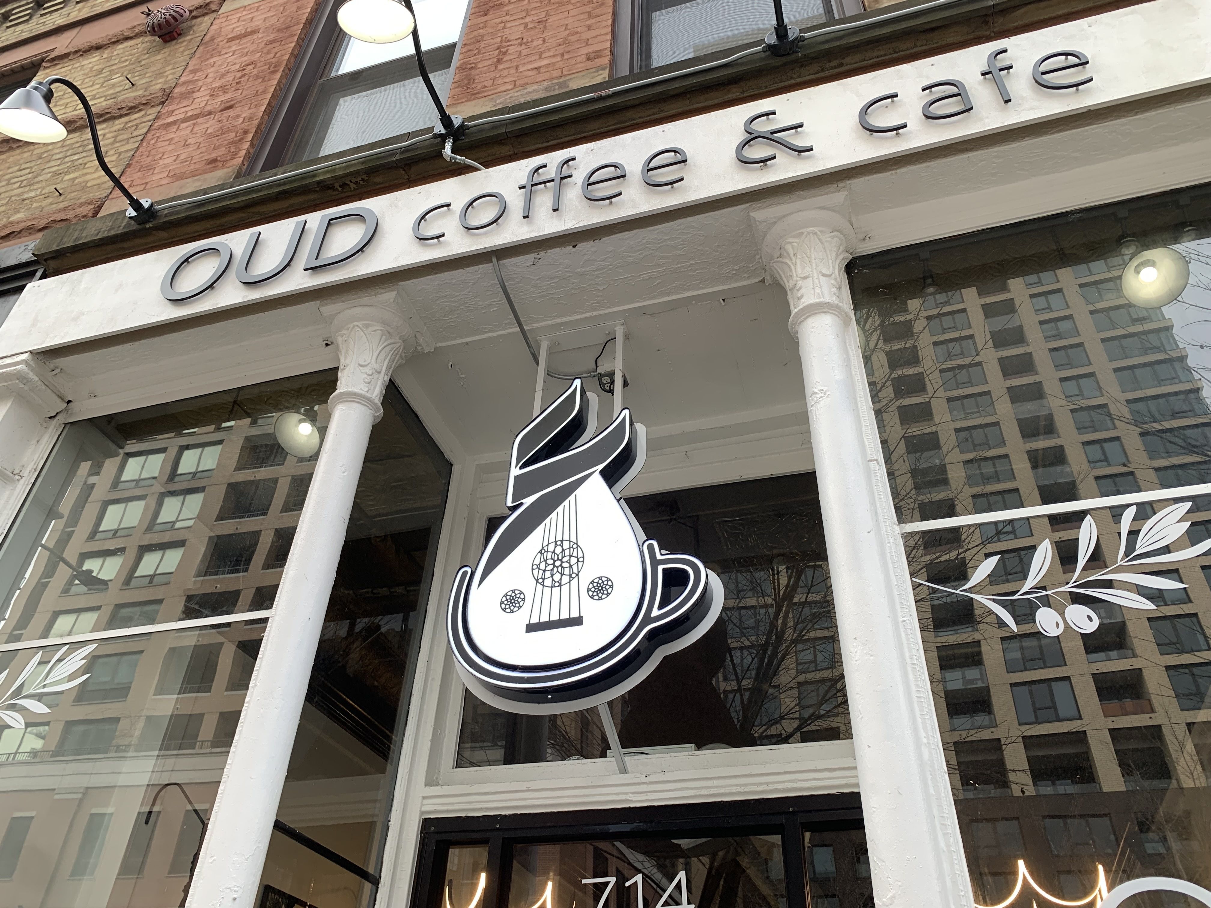 Exterior of OUD coffee & cafe with white pillars and a hanging sign combining a coffee cup and a musical oud instrument in black and white.