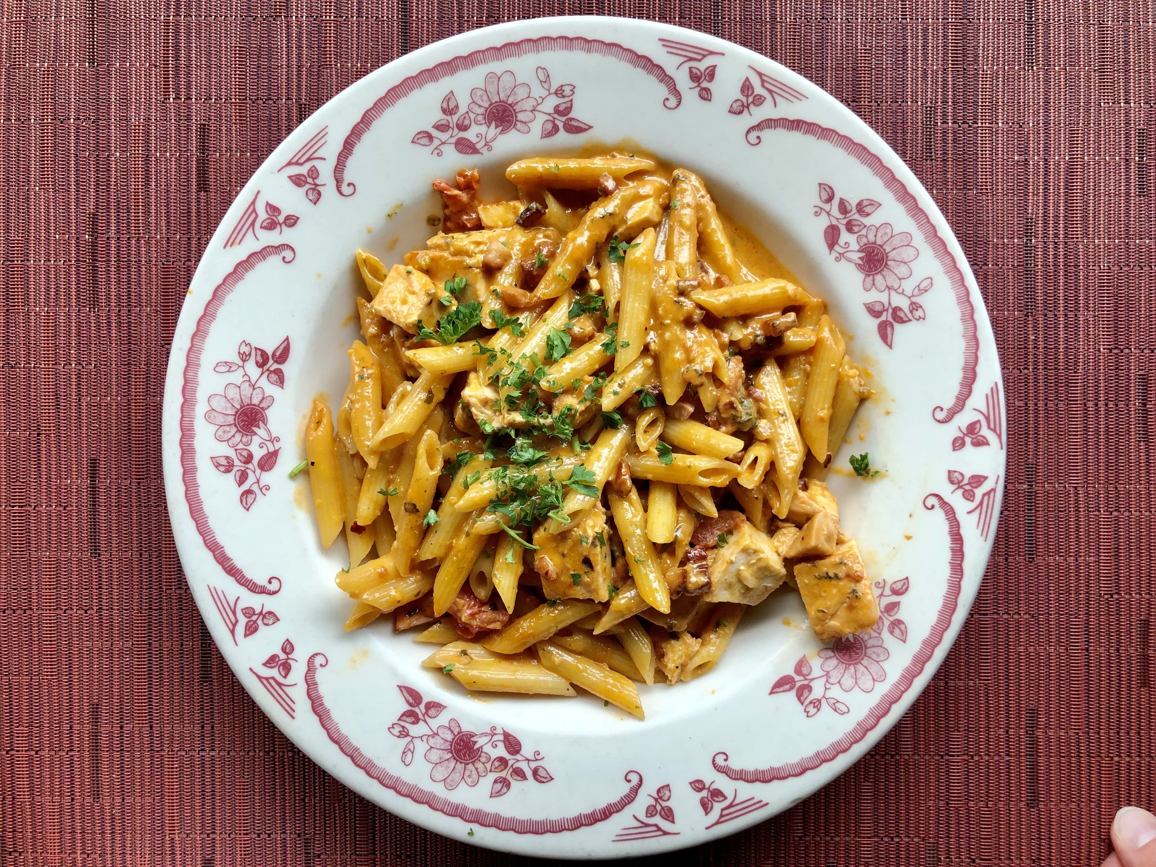A plate of penne pasta in a creamy tomato sauce, garnished with chopped parsley and pieces of chicken, served on a white plate with a red floral border, placed on a red textured tablemat.