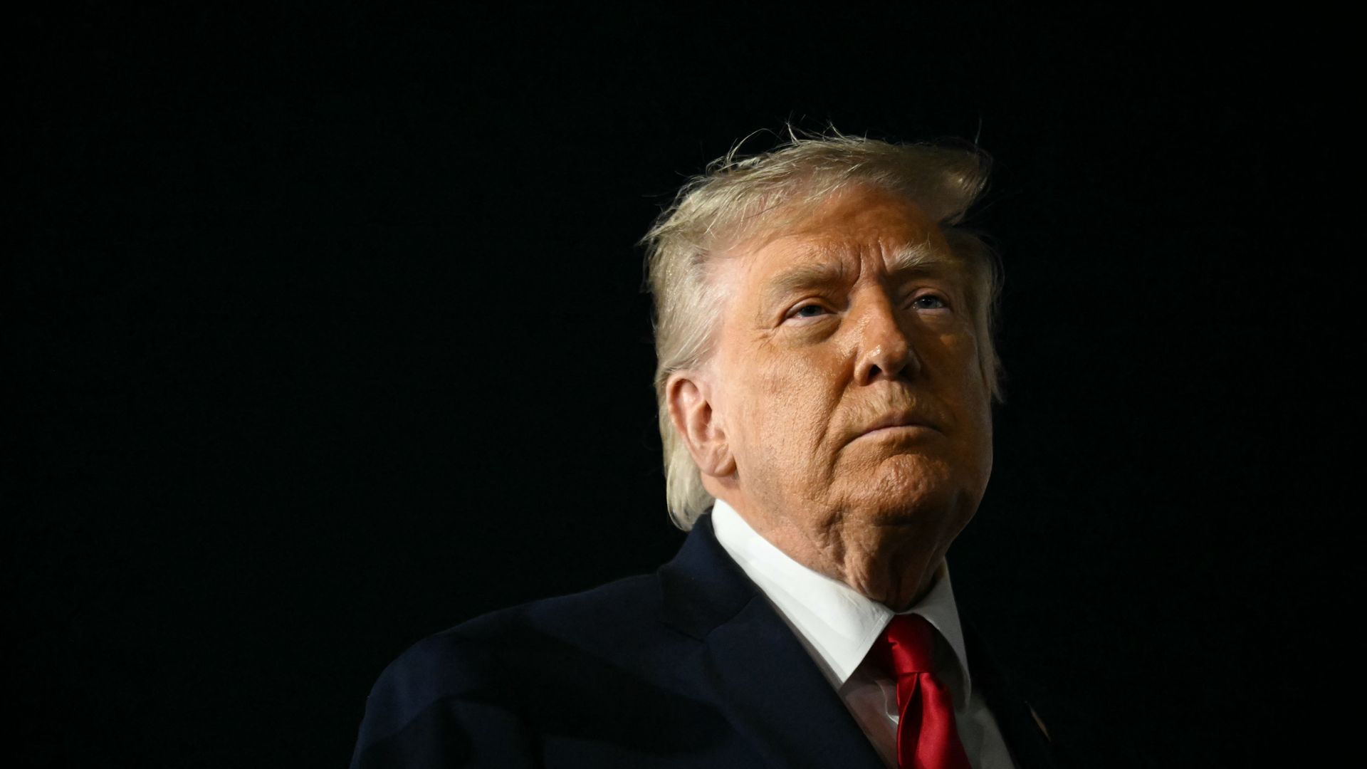 President Donald Trump, wearing a dark suit and red tie, stands against a dark background before boarding Air Force One at JFK Airport in New York.