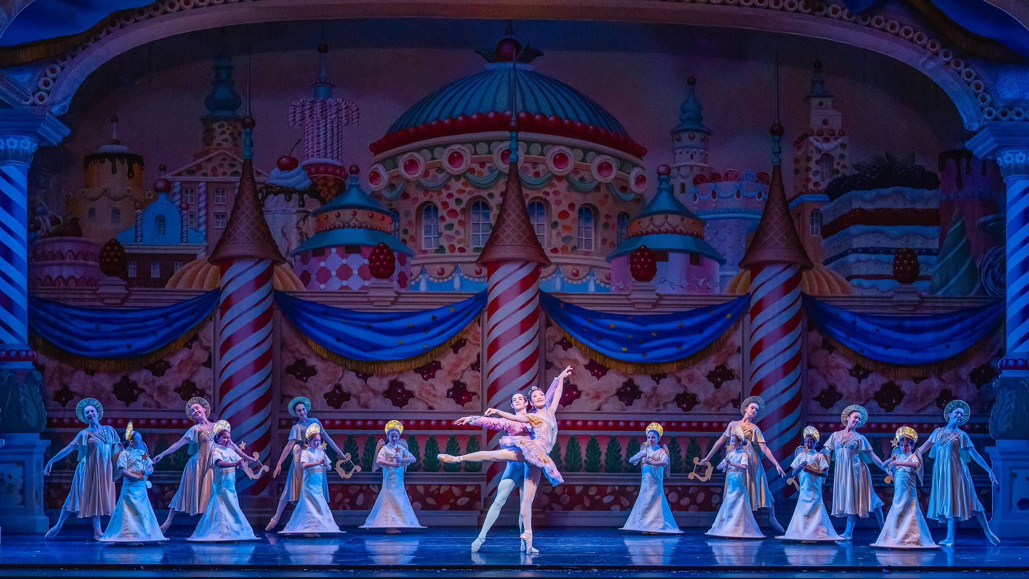 Ballet dancers performing on stage with colorful candy-themed backdrop, including striped pillars, blue curtains, and cake decorations, in elegant costumes under blue lighting.