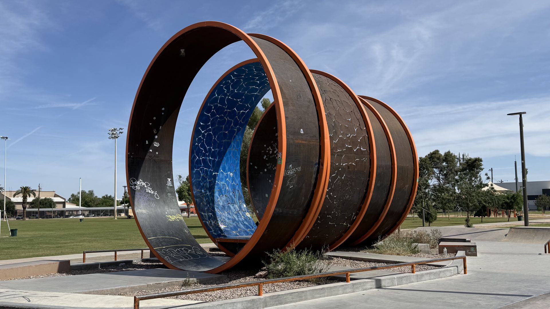 Public art sculpture of large rust-orange circular rings spiraling together, with blue mosaic inner panels and black surfaces, set in a sunny park with grass, trees, and street lamps.