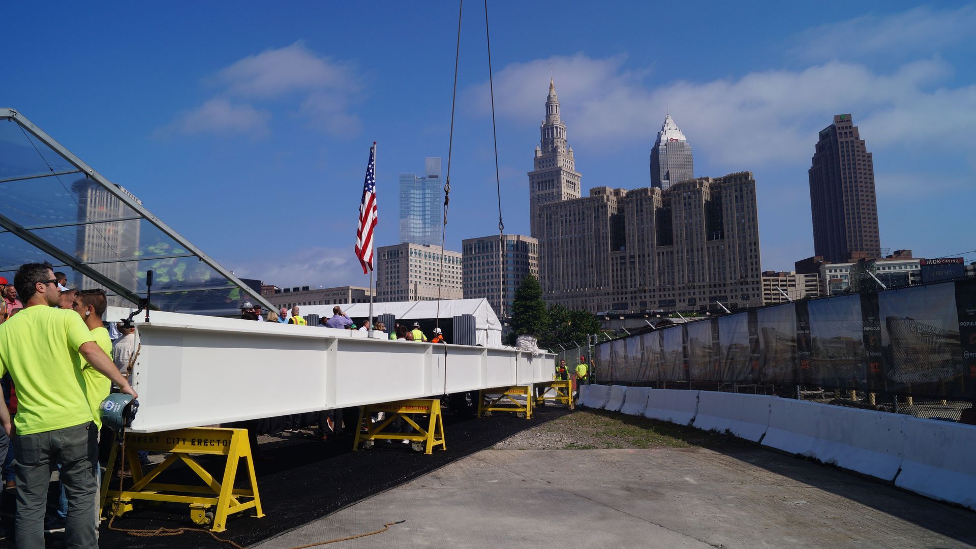 Construction site on a city waterfront with a large white platform and yellow supports. Neon-shirted workers stand near the edge as an American flag waves; tall skyscrapers rise under a blue sky.