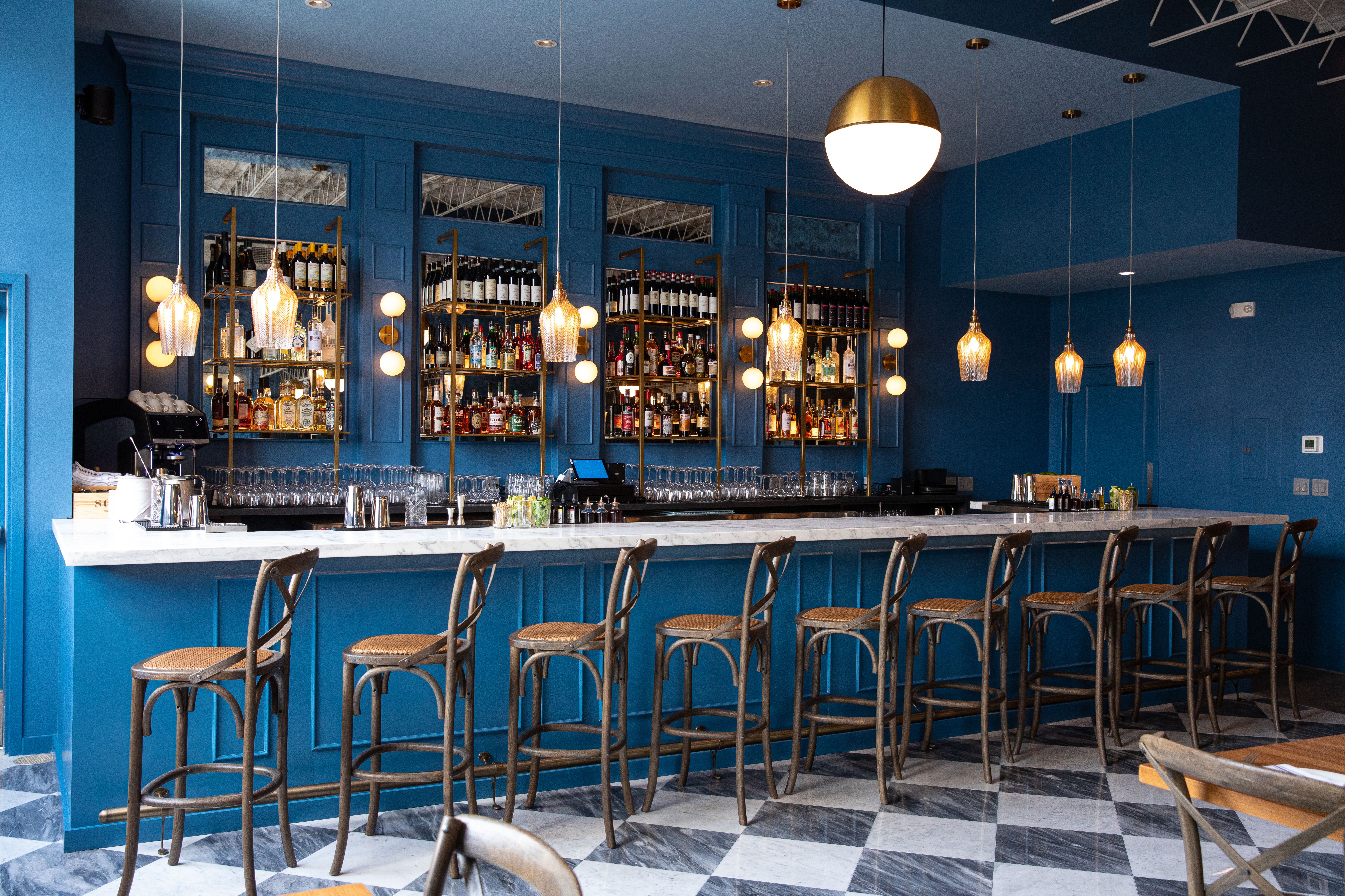A view of the bar inside Osteria Lupo. The walls and bar back are a deep blue, and bronze pendant lights hang from the ceiling. Tall bar chairs await customers.